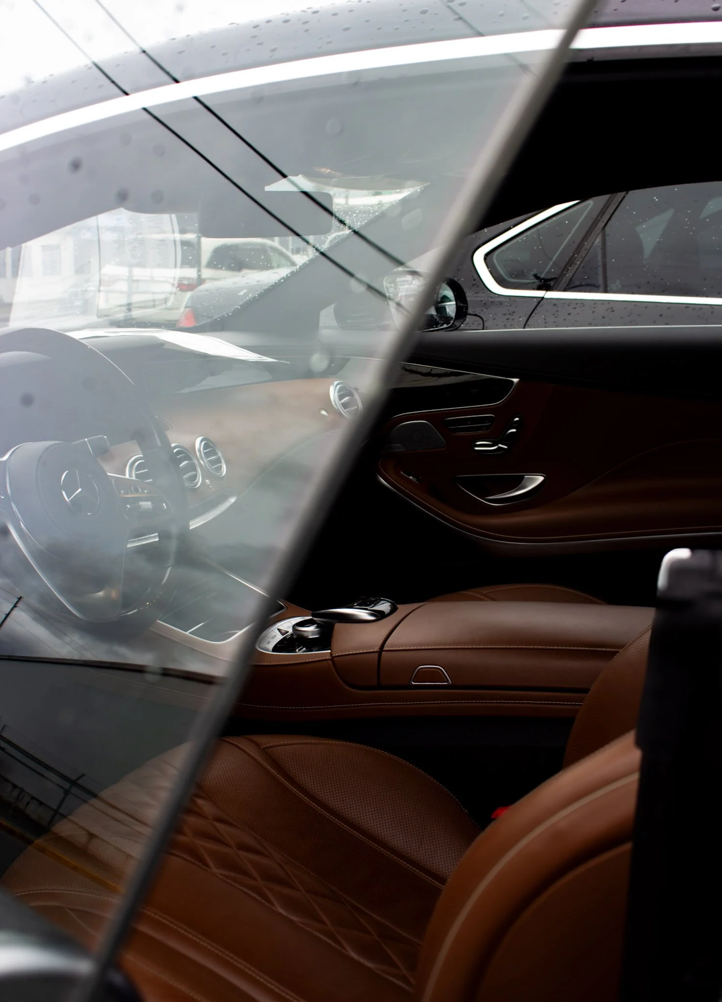 The interior of a luxury car showing brown leather seats, a dashboard, and a visible side window. The photo is taken from outside through the windshield, with some reflections and raindrops visible.