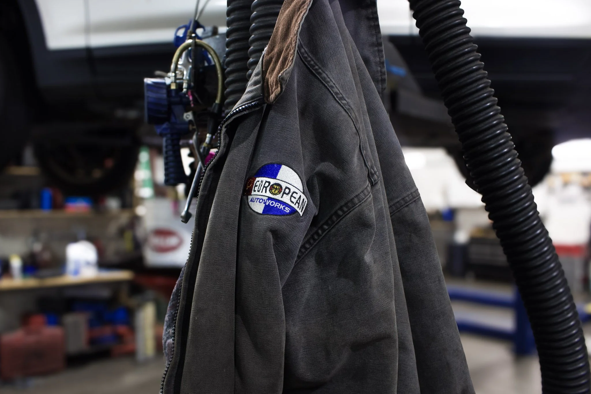 A dark gray work jacket hanging on a hook in an auto repair shop, with a patch reading "European Autoworks" on the sleeve, surrounded by hoses and tools.