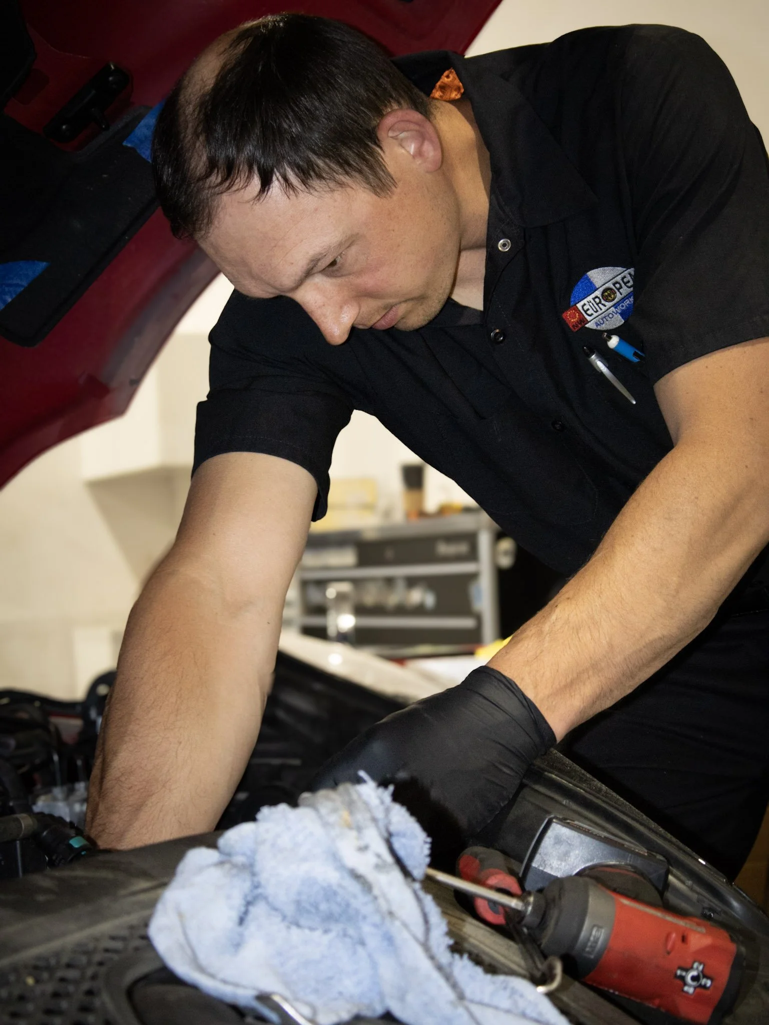A man working on a motorcycle engine, wearing a black shirt with a logo, surrounded by tools and equipment in a garage or workshop.