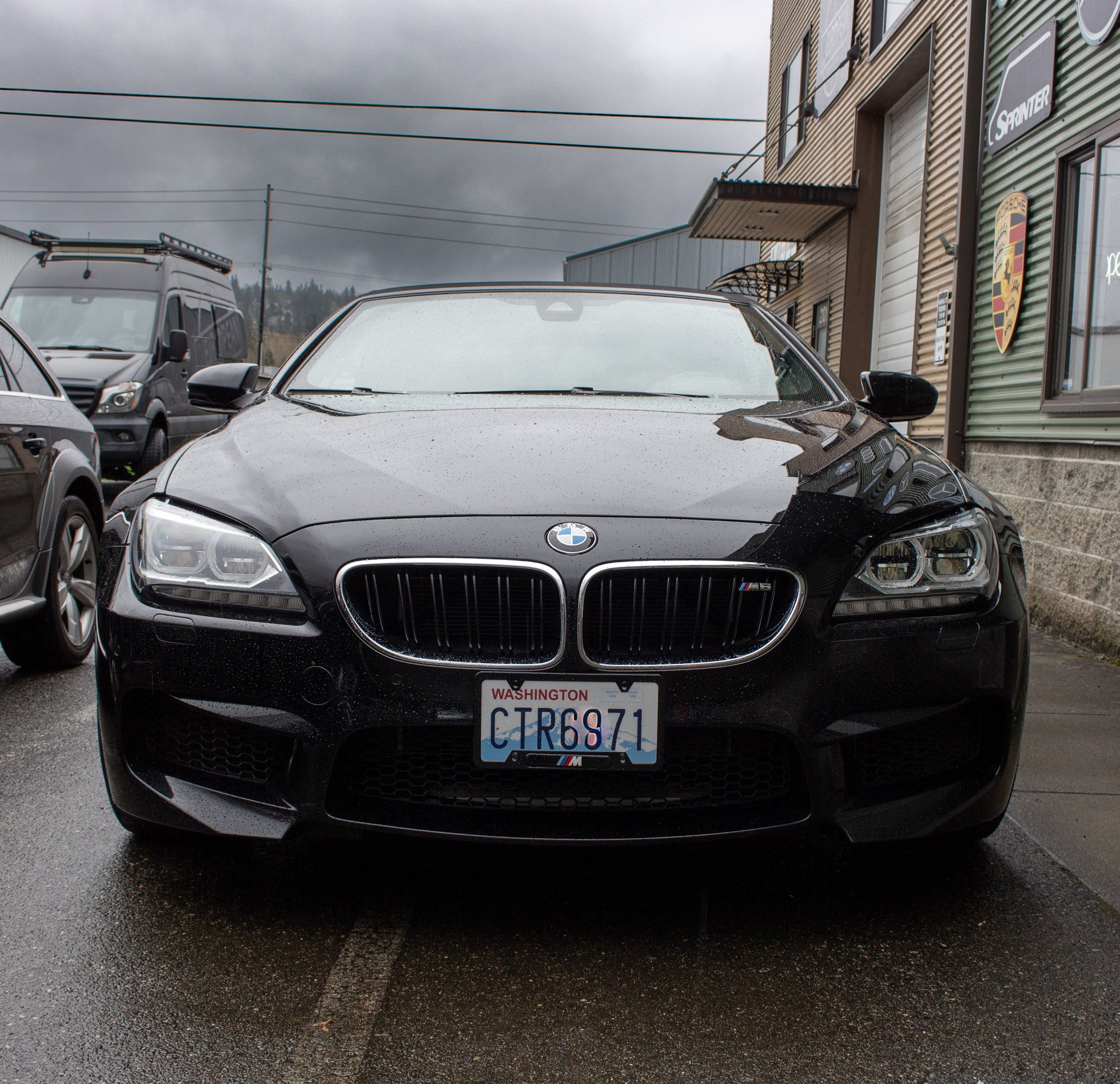 Black BMW car parked outside a building under a cloudy sky, with other vehicles nearby and utility wires overhead.