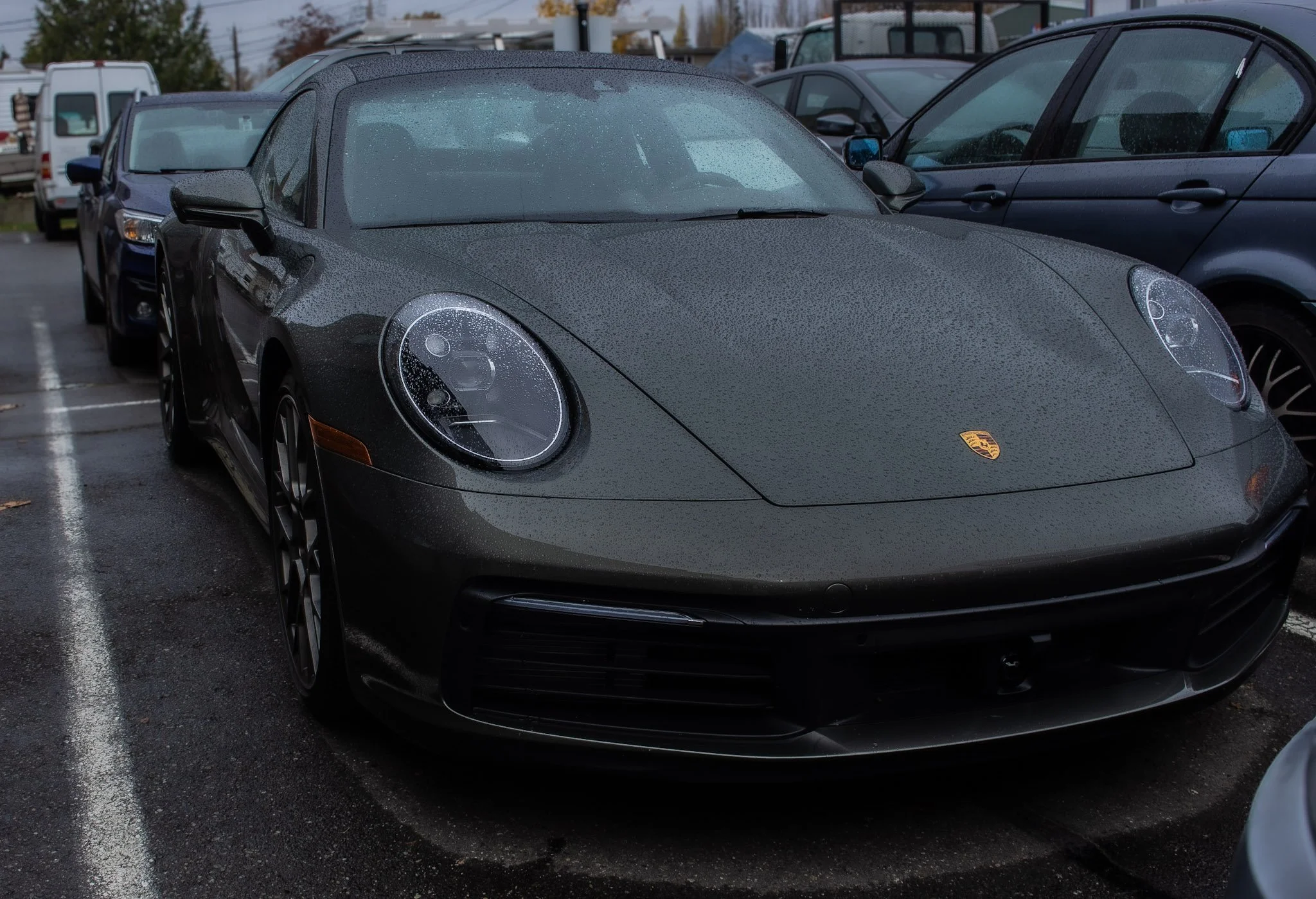 A green Porsche sports car parked in a parking lot with water droplets on its surface, alongside other vehicles.