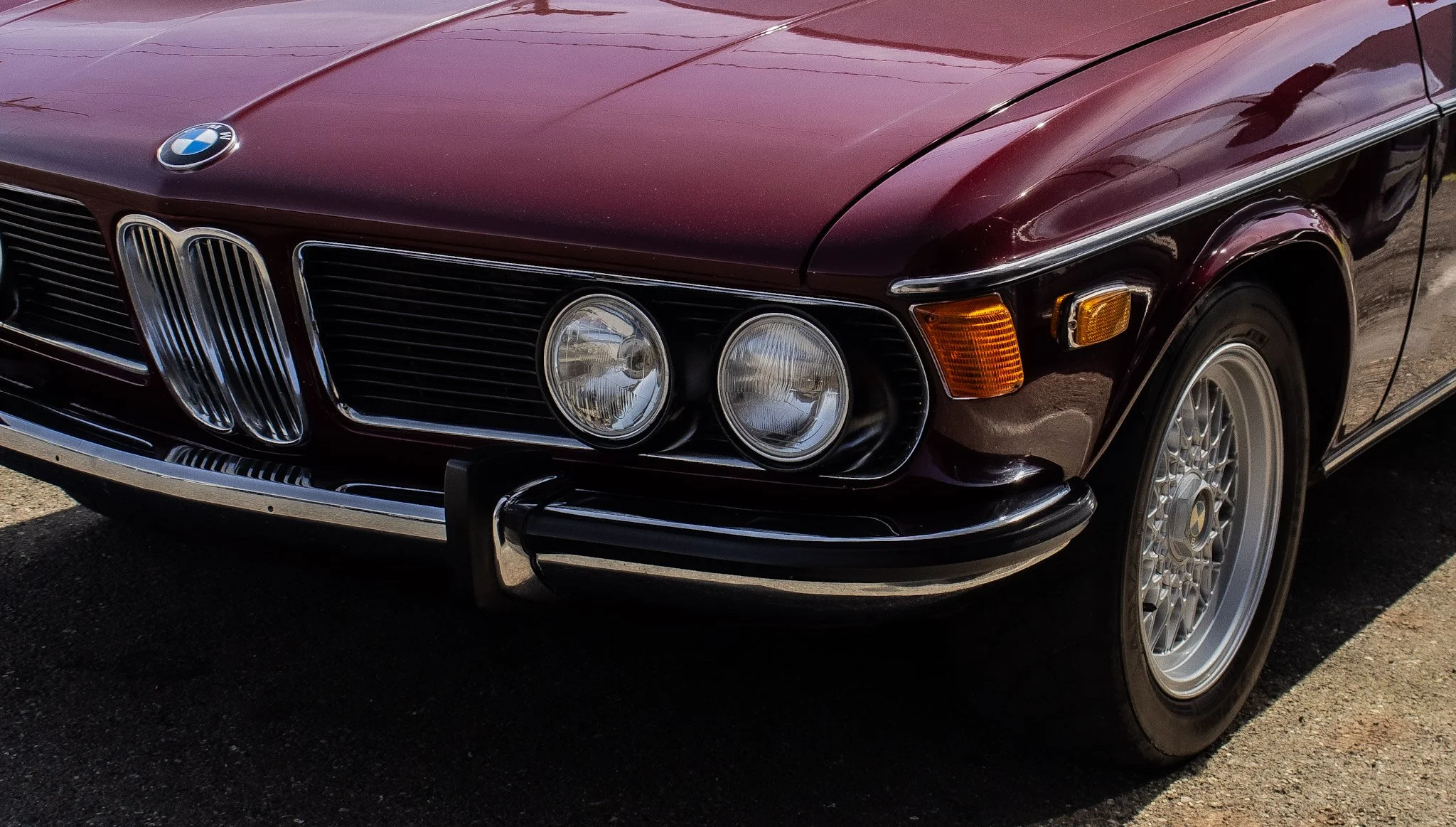 Close-up of a vintage burgundy BMW car with dual round headlights, a kidney grille, and intricate alloy wheels.
