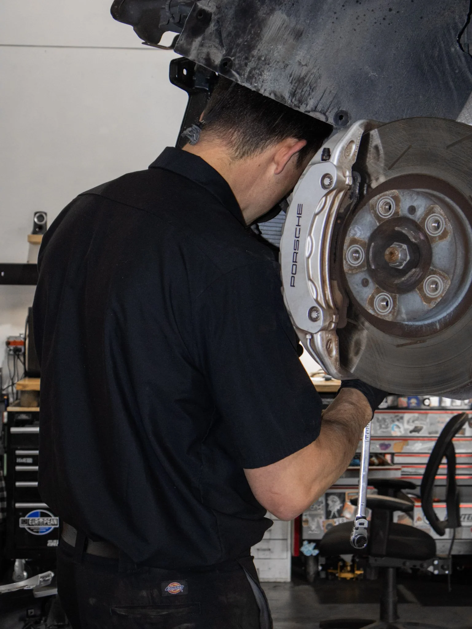 A mechanic in a black shirt working on a car's front brake assembly with a large brake caliper marked 'Porsche.' The mechanic's head is bent down, and their arms are positioned beneath the vehicle's wheel well in a garage or workshop.