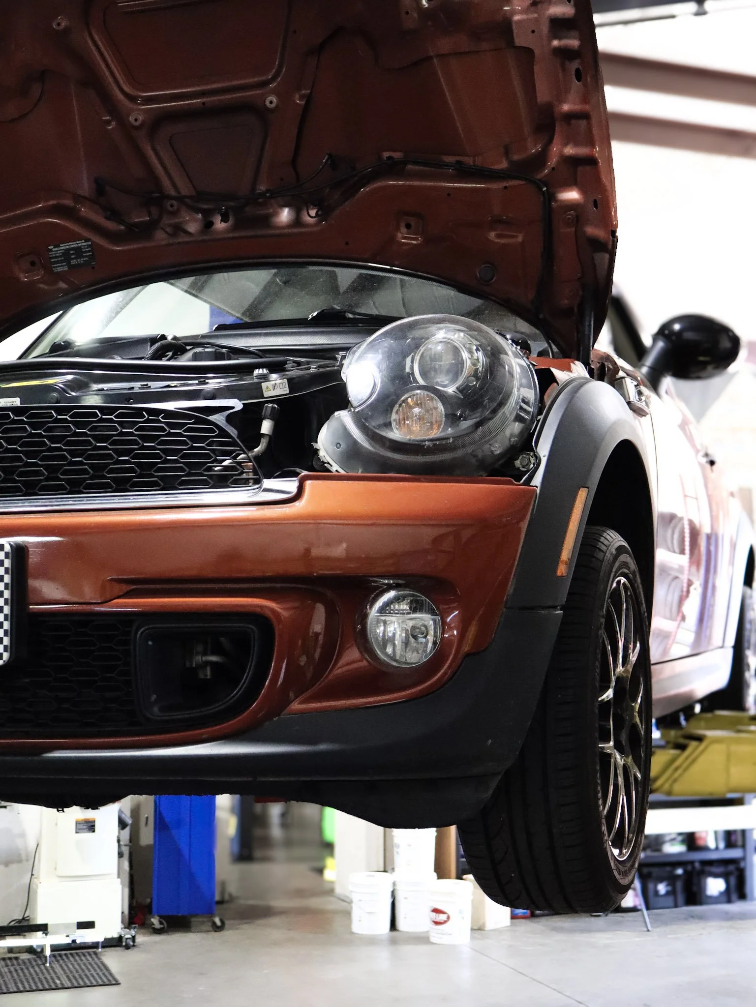 A red car is lifted on a hydraulic lift in a garage, with the hood open. The garage has various tools, buckets, and equipment visible in the background.