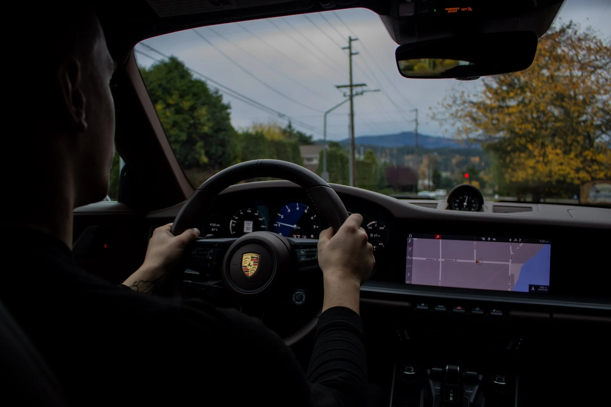 Interior of a Porsche car with a driver holding the steering wheel, dashboard gauges, and a GPS navigation screen, during daytime with trees and power lines outside.