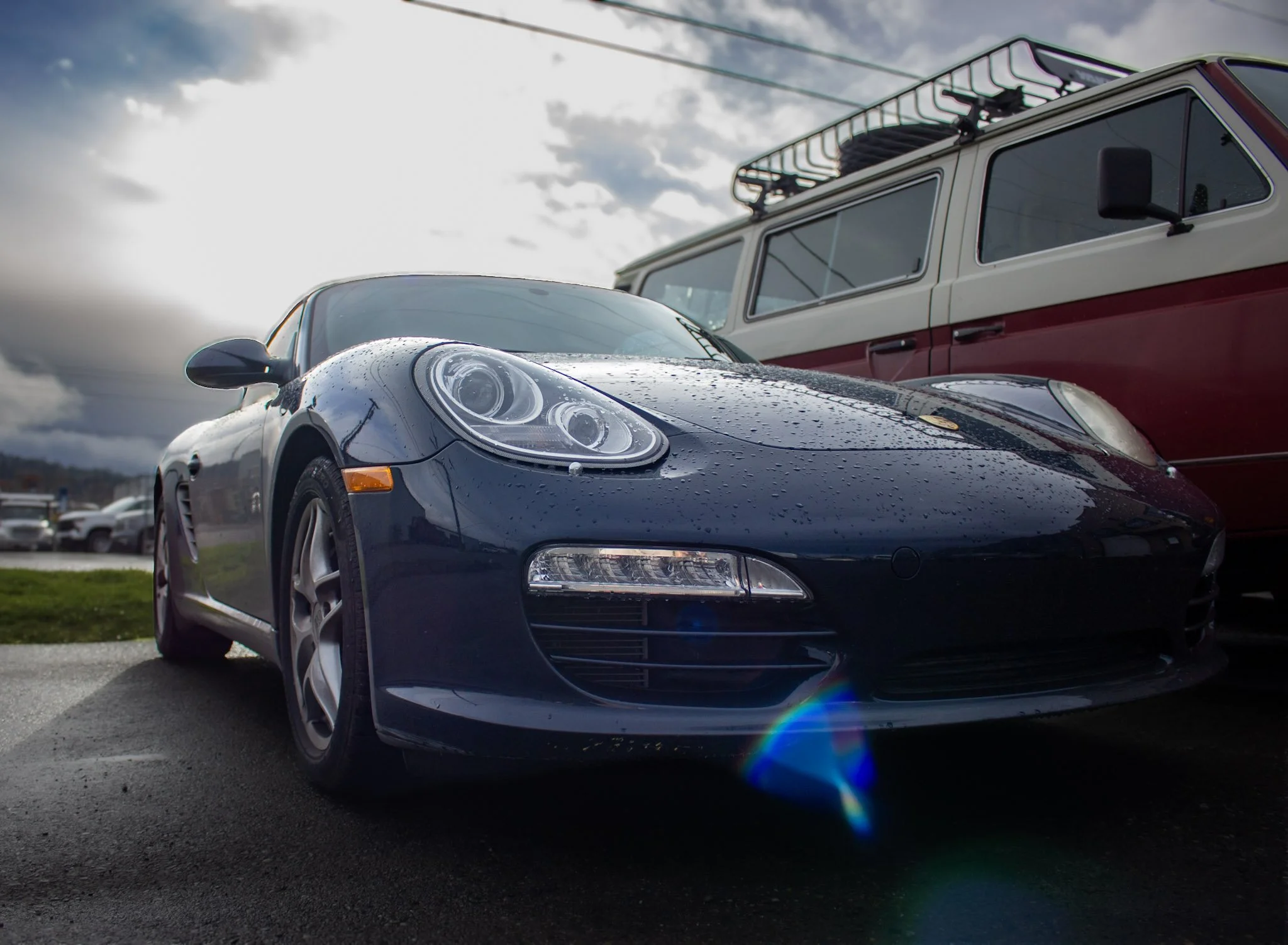 A black Porsche sports car with raindrops on its surface parked outdoors next to a red and white vintage van under a cloudy sky.