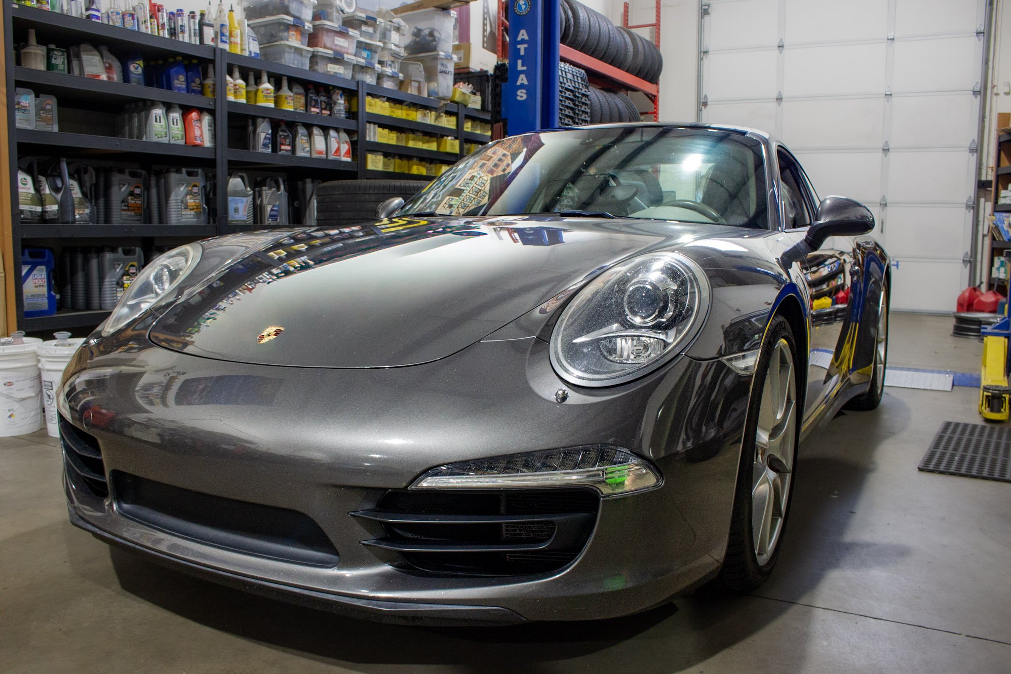 A gray Porsche sports car parked inside a garage or workshop, with shelves of automotive supplies and tires in the background.