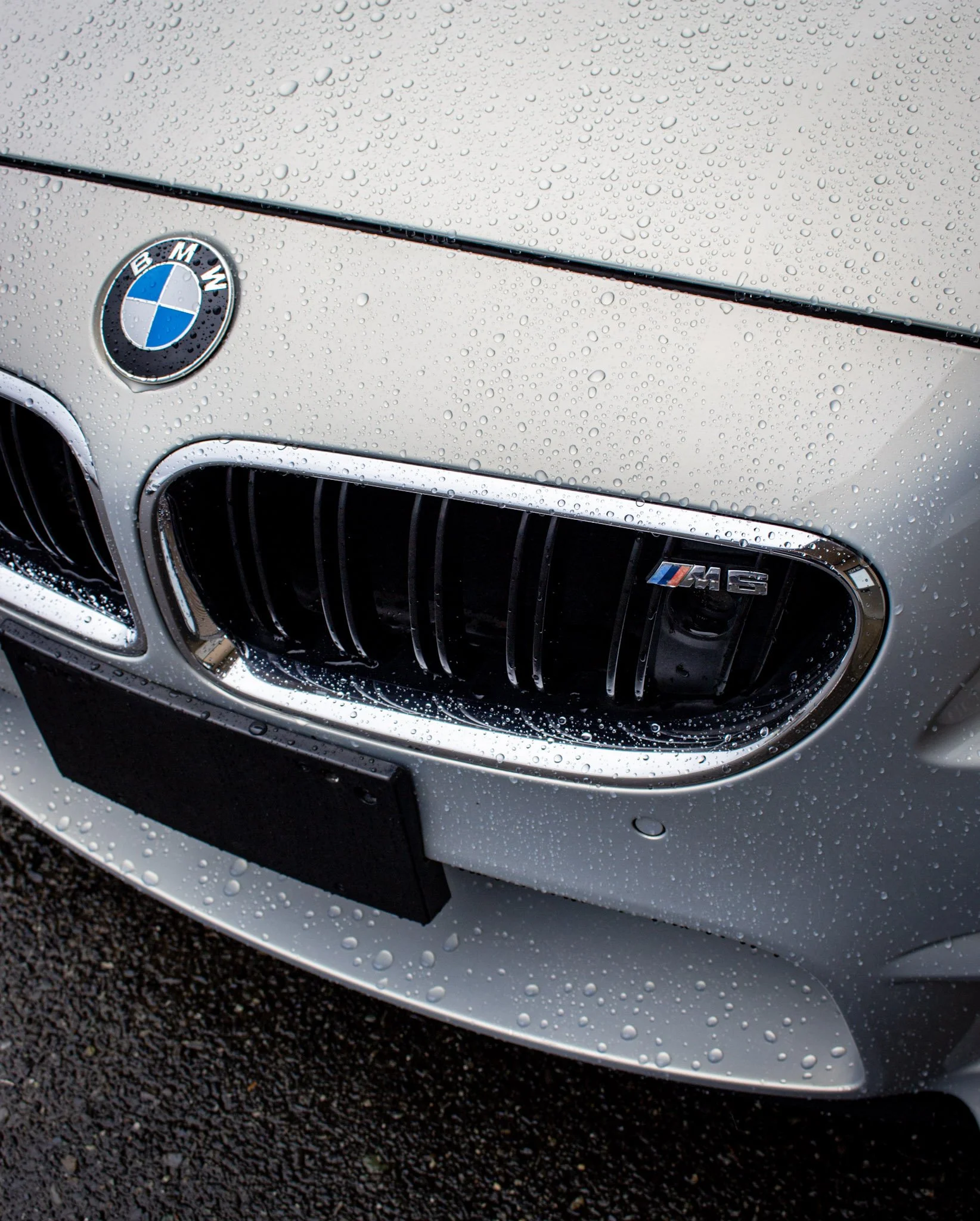 Close-up of the front of a white BMW M2 sports car, showing the BMW emblem on the hood, water droplets on the surface, and part of the front grille and bumper.