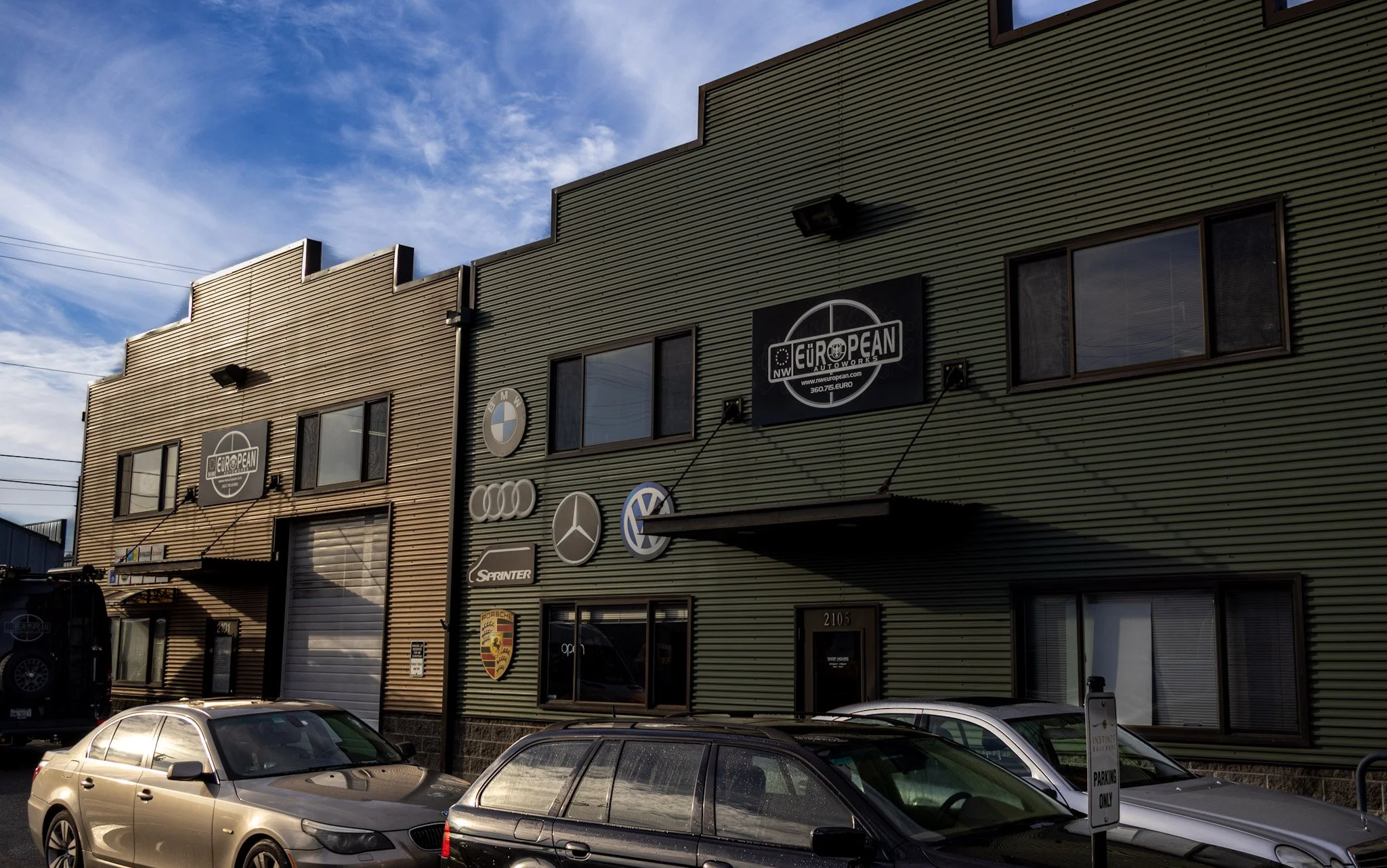 Exterior of a commercial building with logos of European car brands such as BMW, Mercedes-Benz, Audi, Porsche, Volkswagen, and Sprinter on the wall, along with parked cars in front.