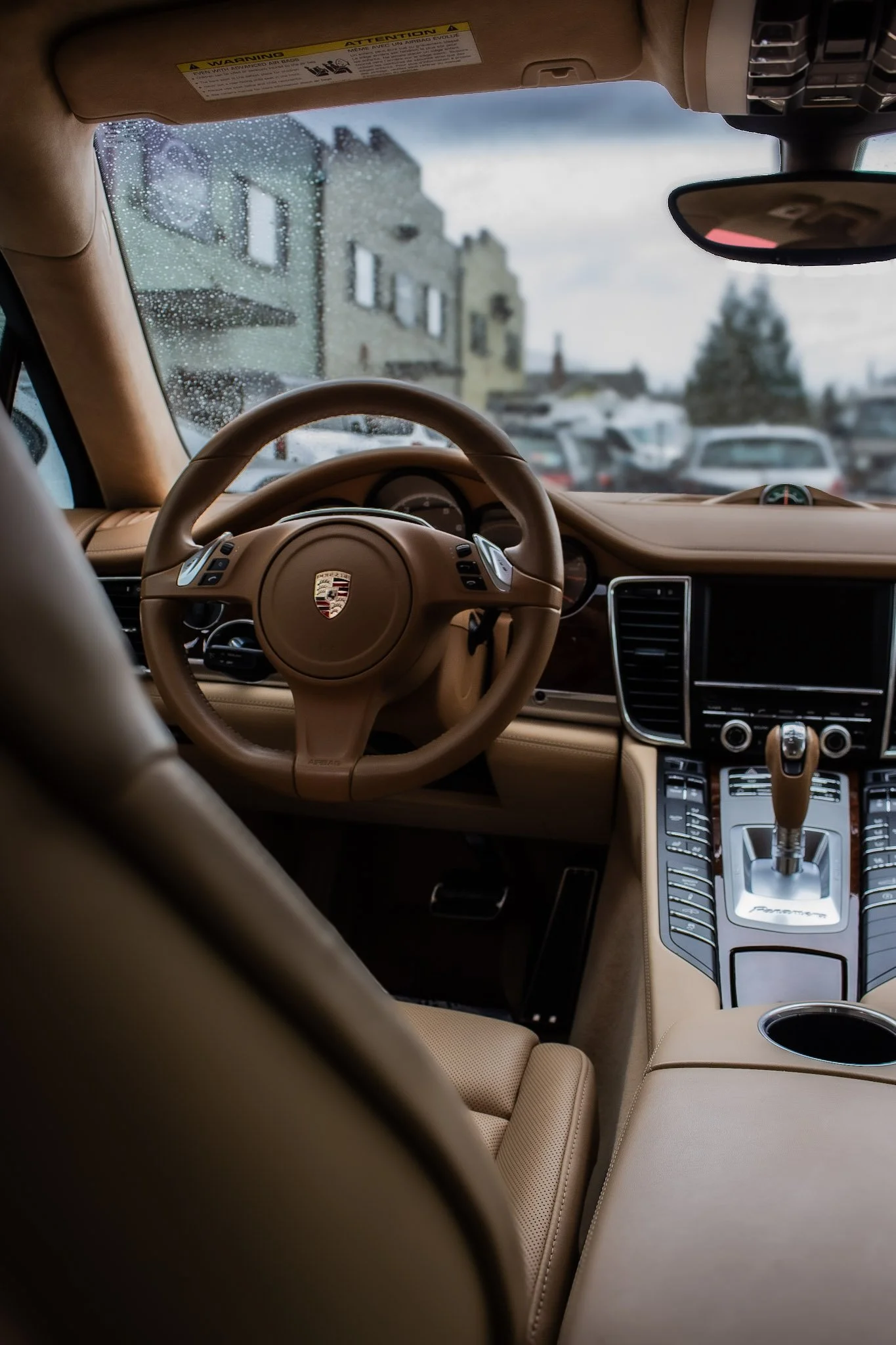 View from inside a luxury car with a tan leather interior, showing the steering wheel with Porsche emblem, center console, gear shift, and a rearview mirror. Rain droplets are on the front windshield, and a blurry cityscape with parked cars and build