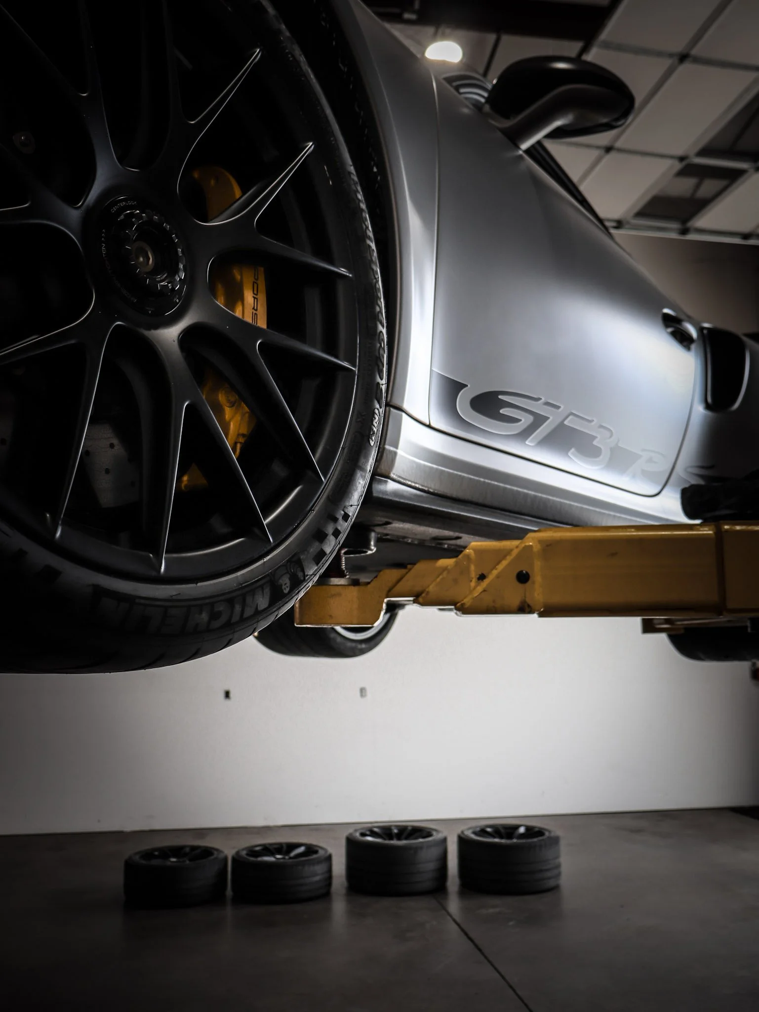 A silver Porsche GT3 car on a hydraulic lift in a garage, with tires stacked on the floor below.