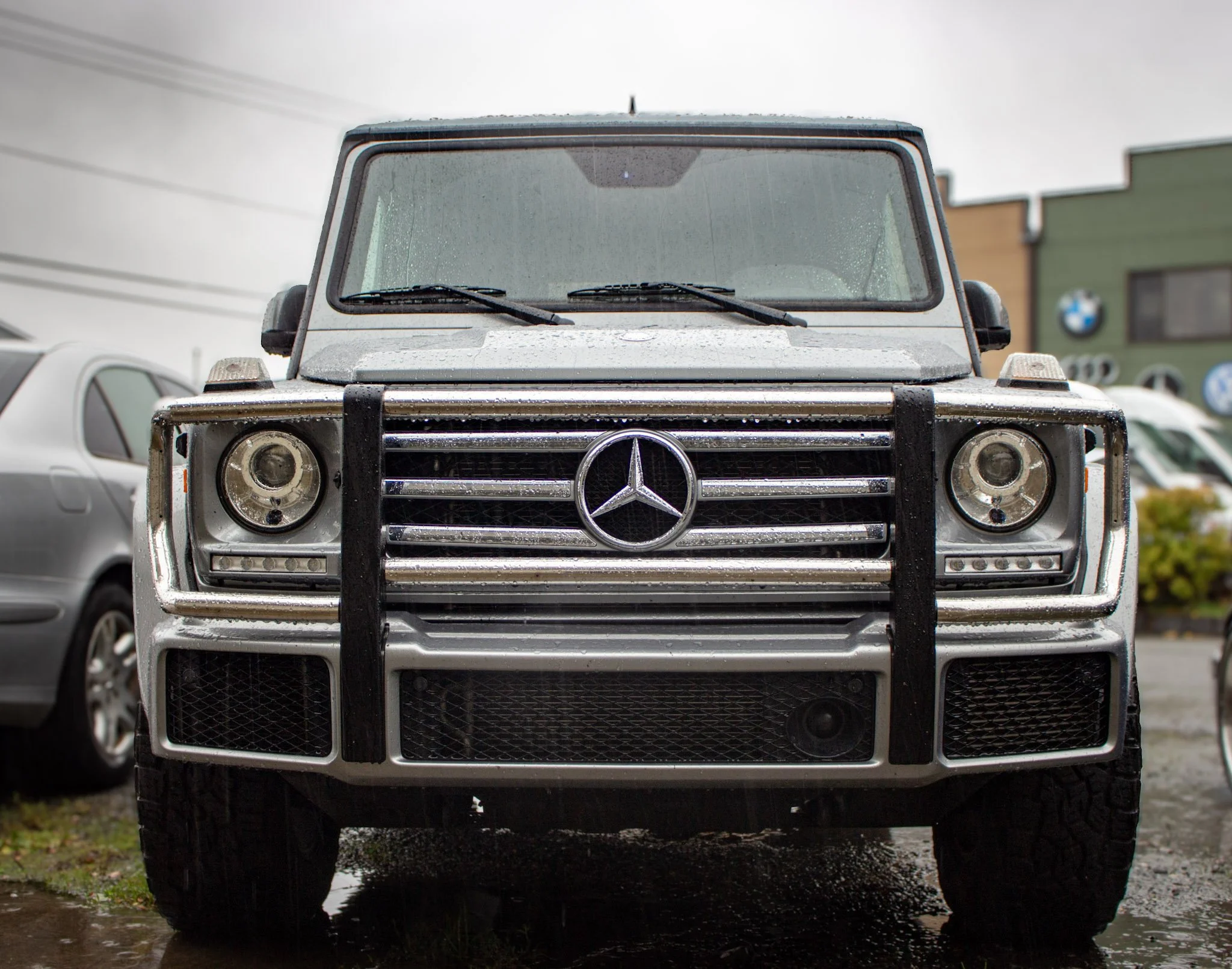 Front view of a Mercedes-Benz G-Class SUV with a black strap guard on the grille, parked with rain droplets visible on the windshield and hood.