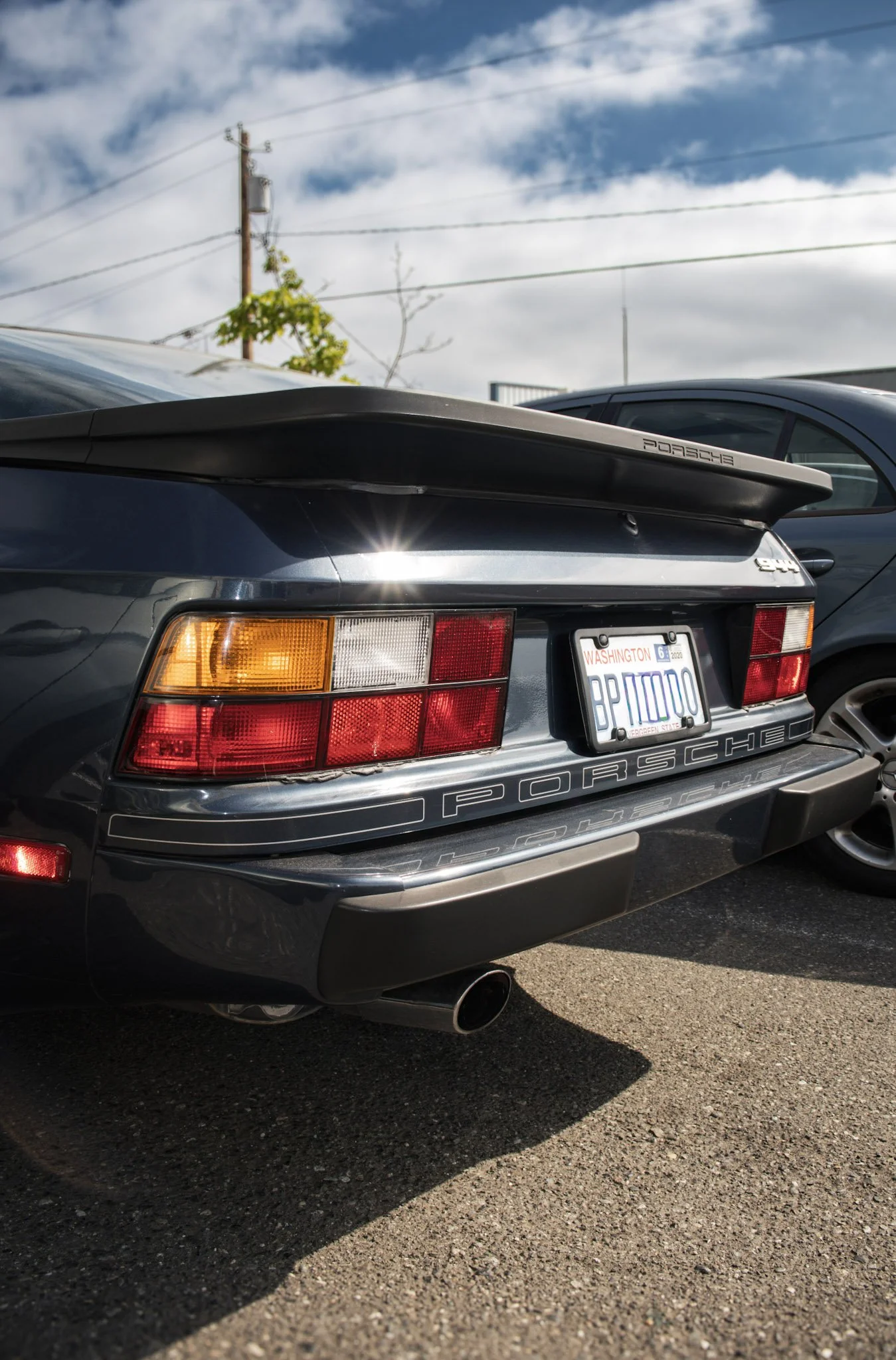 Rear view of a dark gray Porsche 911 with a spoiler, parked on asphalt, with partial view of a silver car beside it, under a partly cloudy sky with power lines and a utility pole in the background.