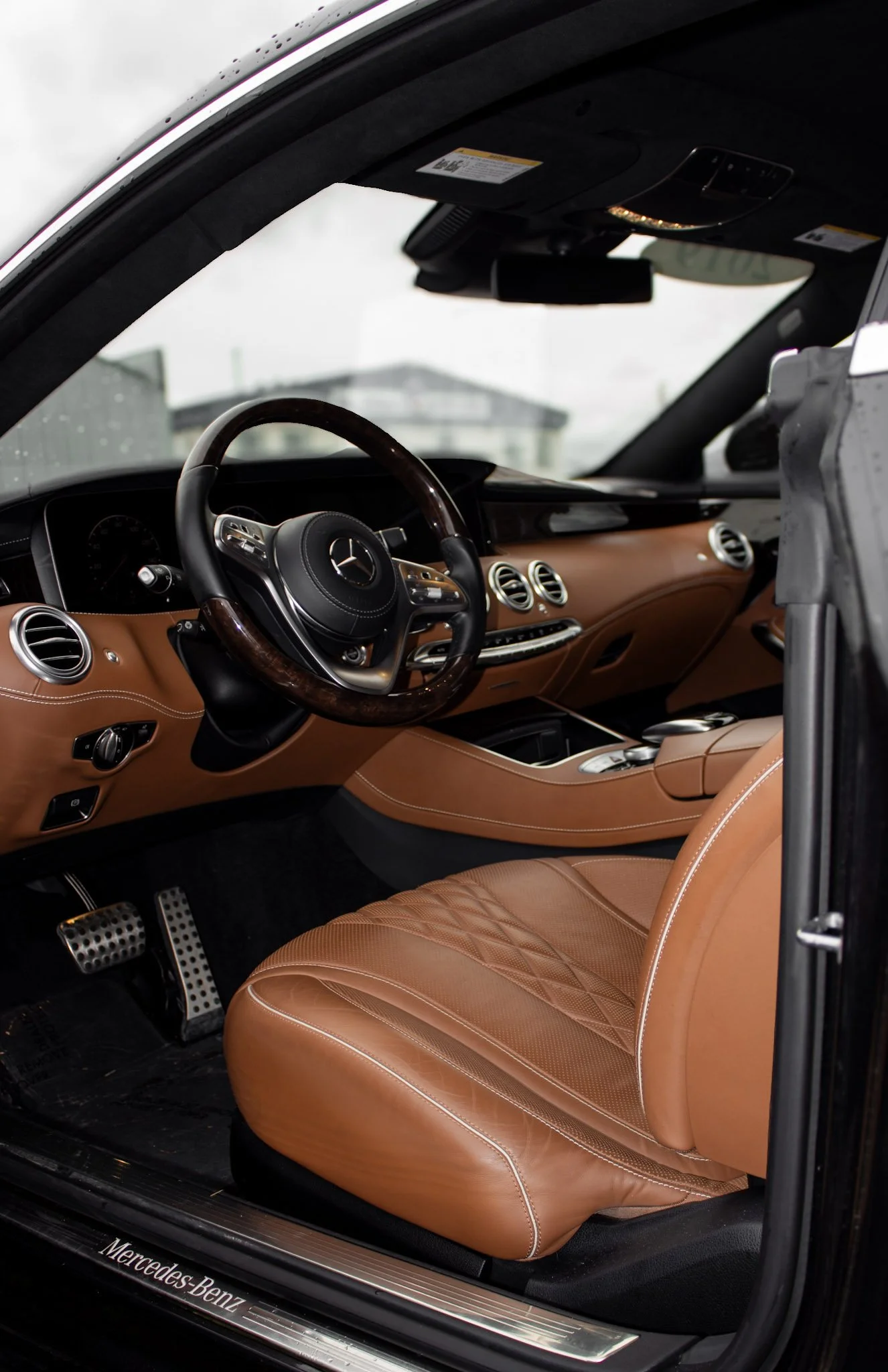 The image shows the interior of a luxury Mercedes-Benz vehicle with tan quilted leather seats, a leather and wood steering wheel, and a dashboard with air vents and controls. The photo is taken from the passenger side with part of the dashboard and t