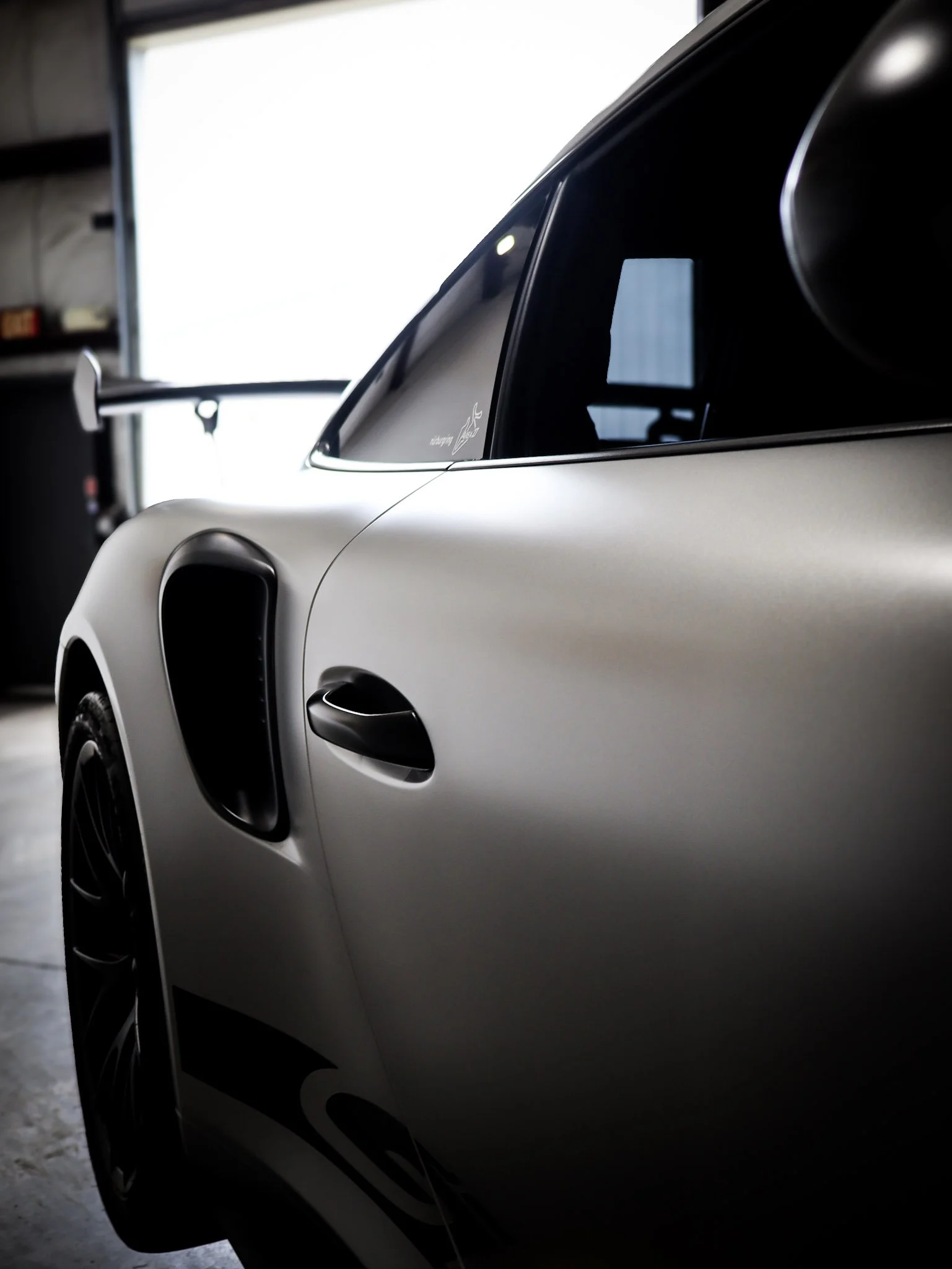 Close-up of a modern electric or hybrid car parked indoors with a background of a partly open garage door and shelves, showing part of the car's side window and door.