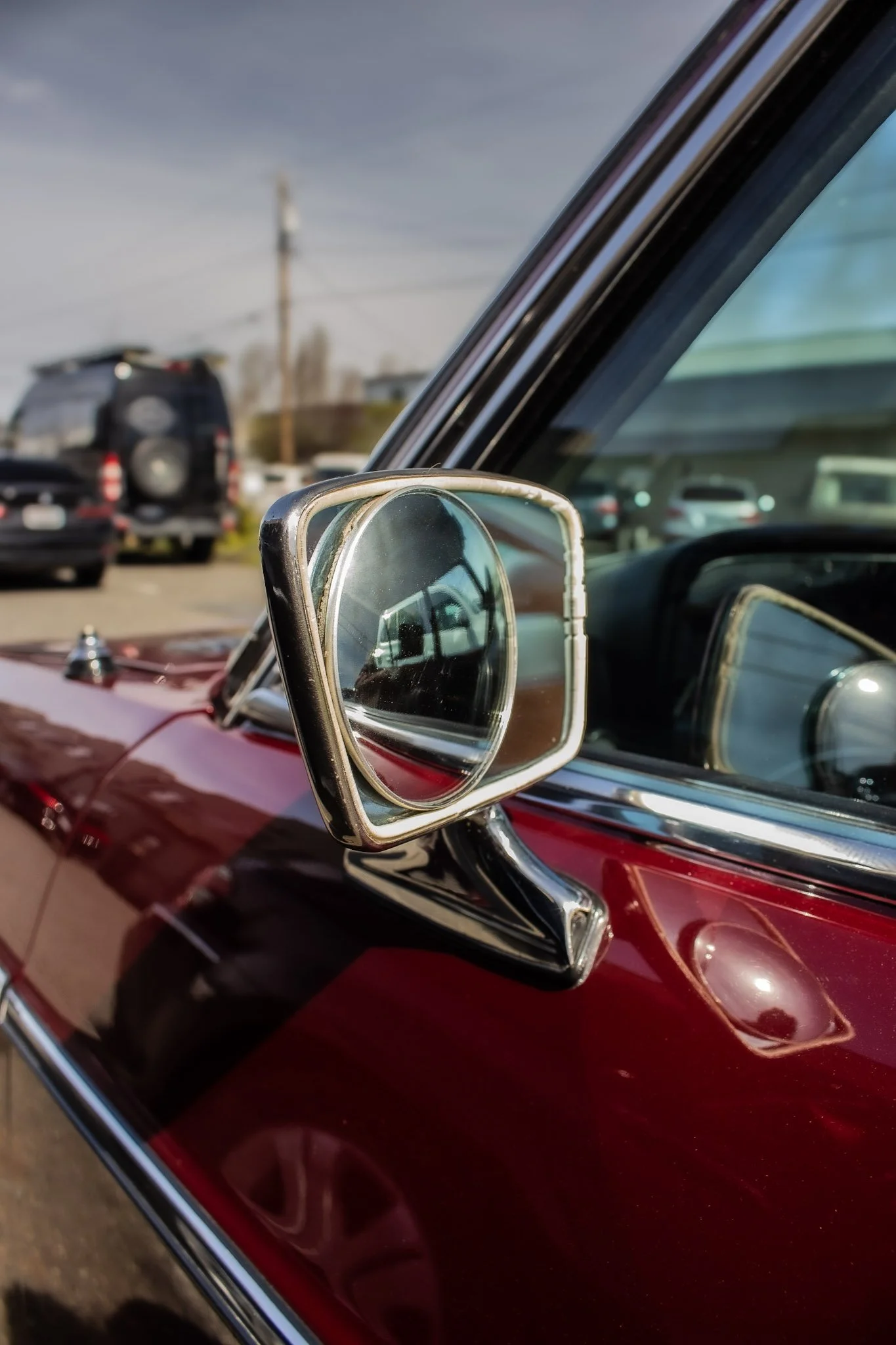 Close-up of a vintage red car's side mirror reflecting part of the car and the parking lot behind it, with other vehicles and a utility pole visible in the background.