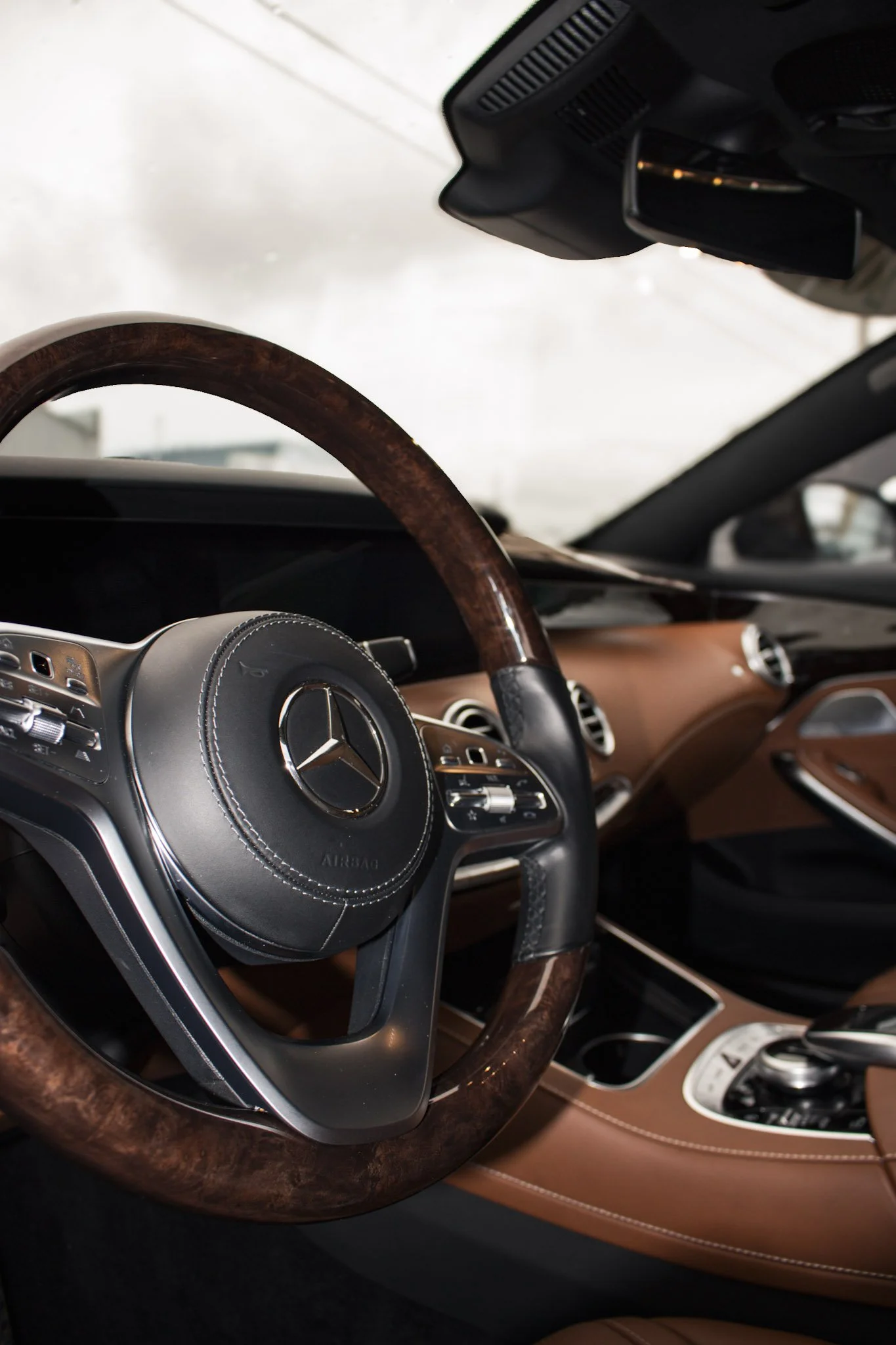 The interior of a luxury car featuring a wooden steering wheel, a Mercedes-Benz logo on the steering hub, dashboard with tree color tones, leather seats, and various control buttons.