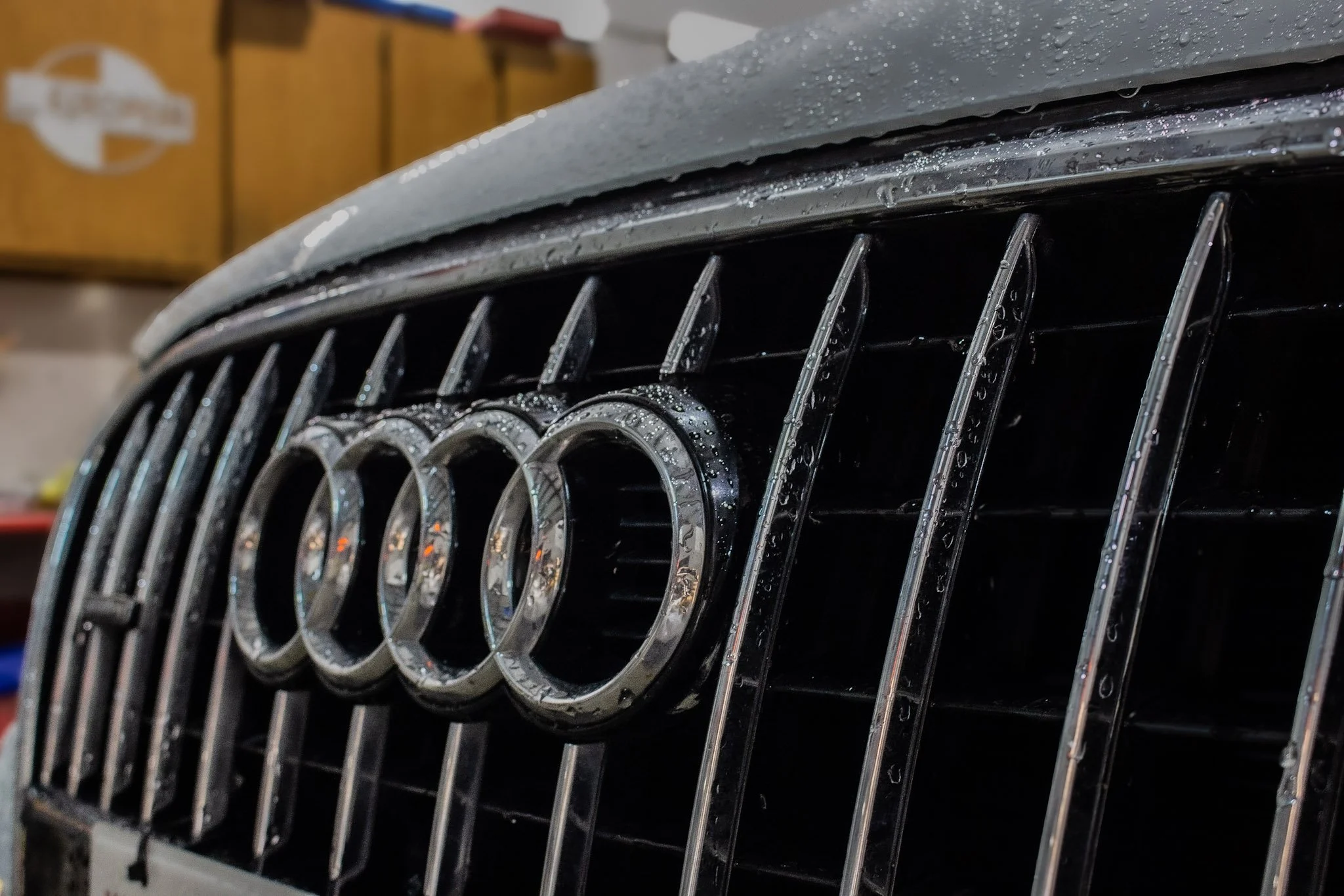 Close-up of a black Audi car grille with the four-ring logo, wet with water droplets, in a garage setting.