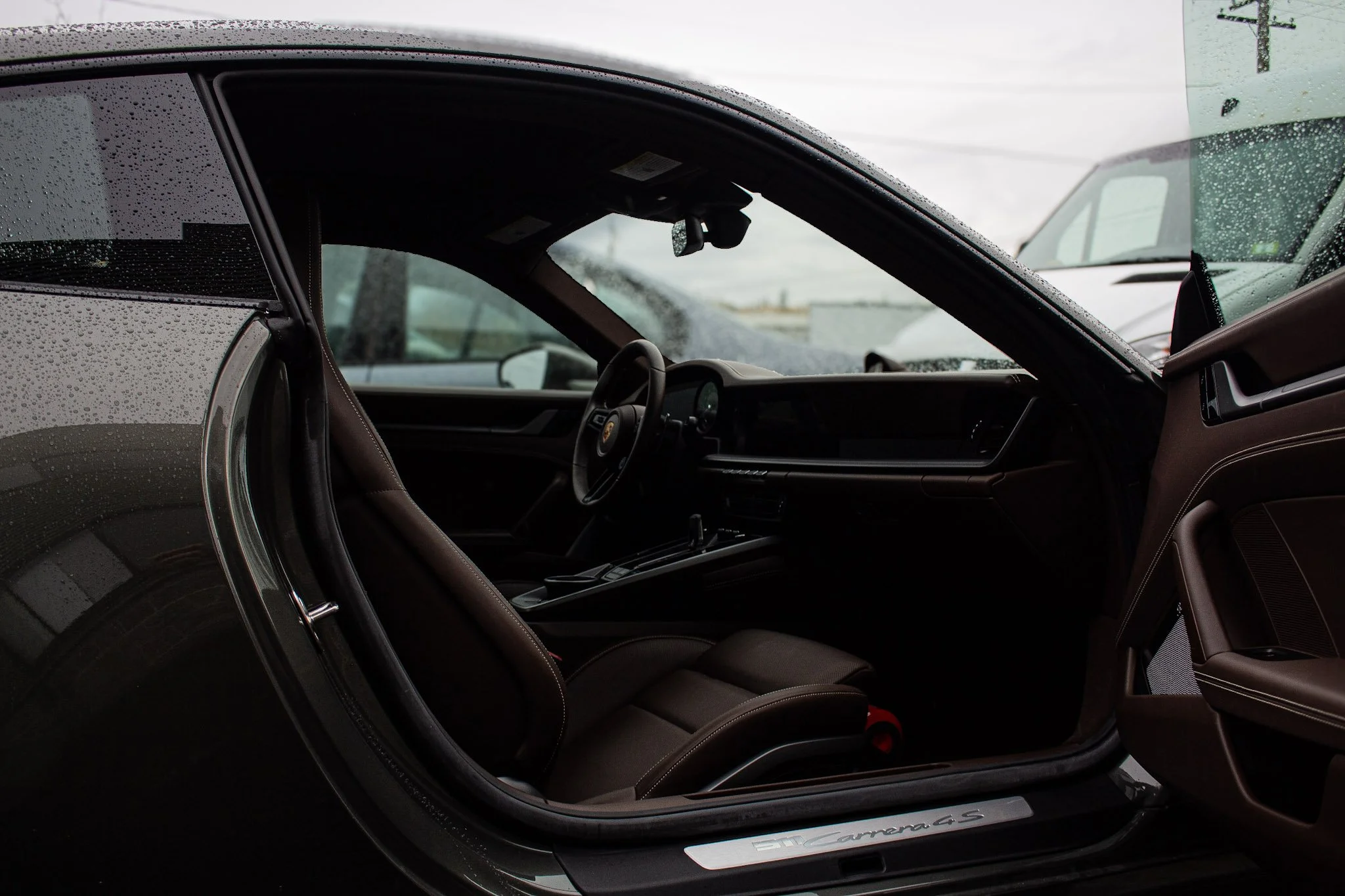 Interior view of a black Porsche 911 Carrera 4 GTS, showing the driver's seat, steering wheel, and dashboard through the open door, with rain droplets on the windshield.