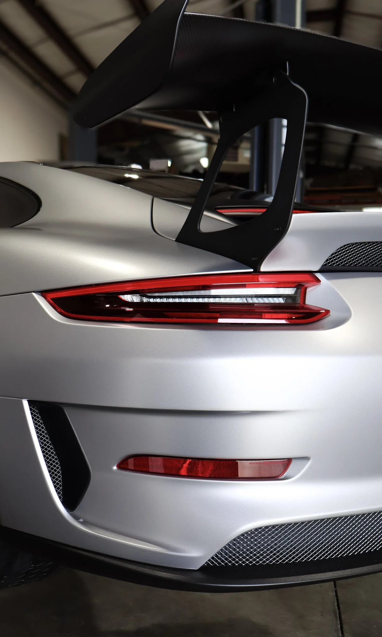 Close-up of the rear section of a silver sports car with a large black rear wing and sleek tail lights, parked indoors.