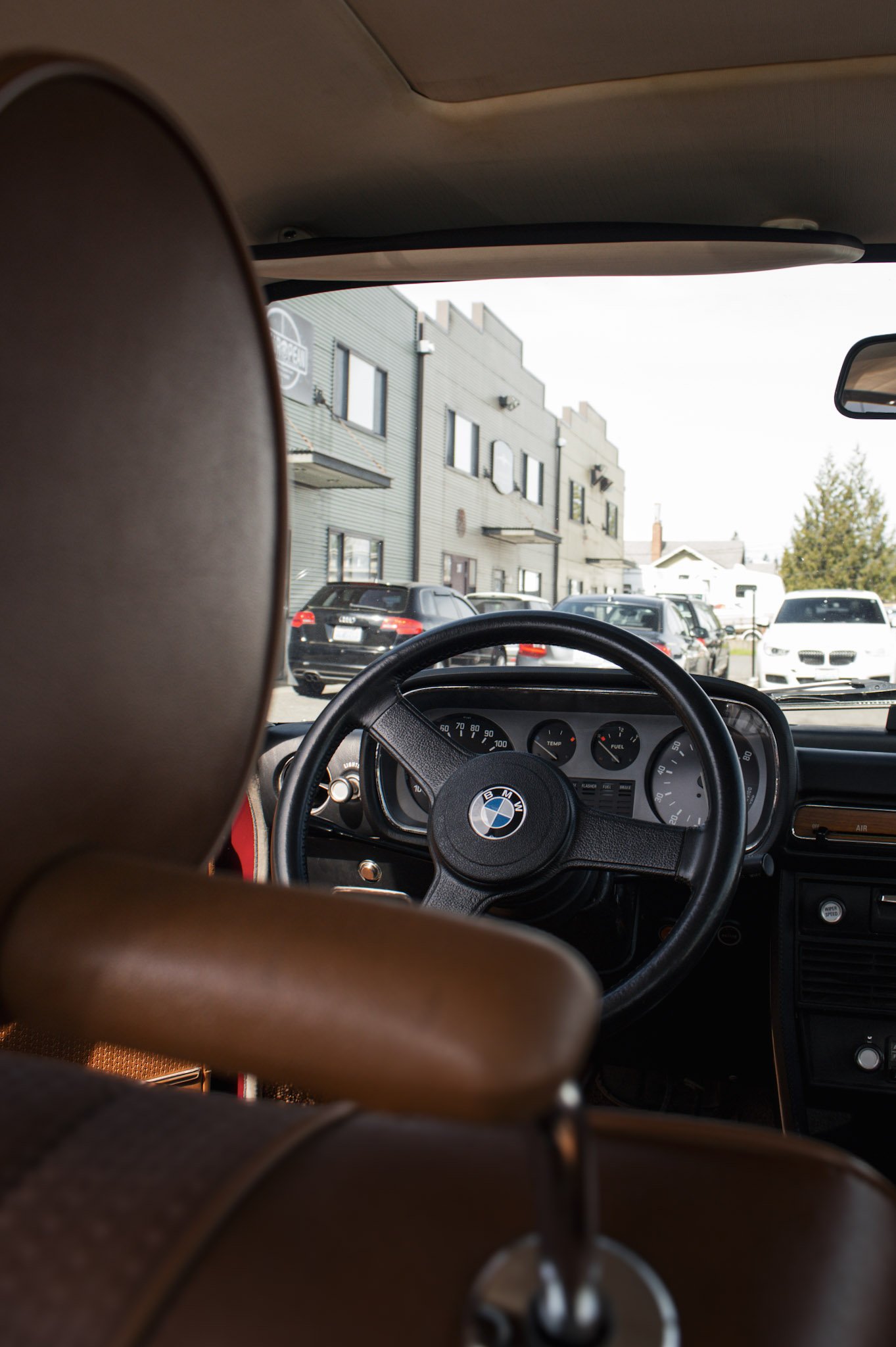Photo taken from inside a car, showing the steering wheel with a BMW logo, the dashboard, and the view outside through the windshield. Outside, there are parked cars and buildings in an urban area.