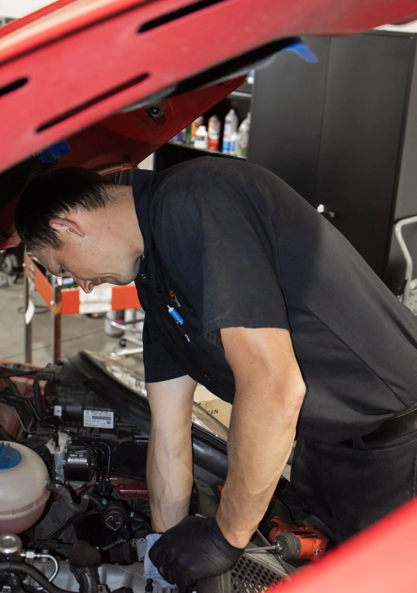 Man working on a car engine in a garage, leaning over the open hood, with shelves of automotive supplies in the background.