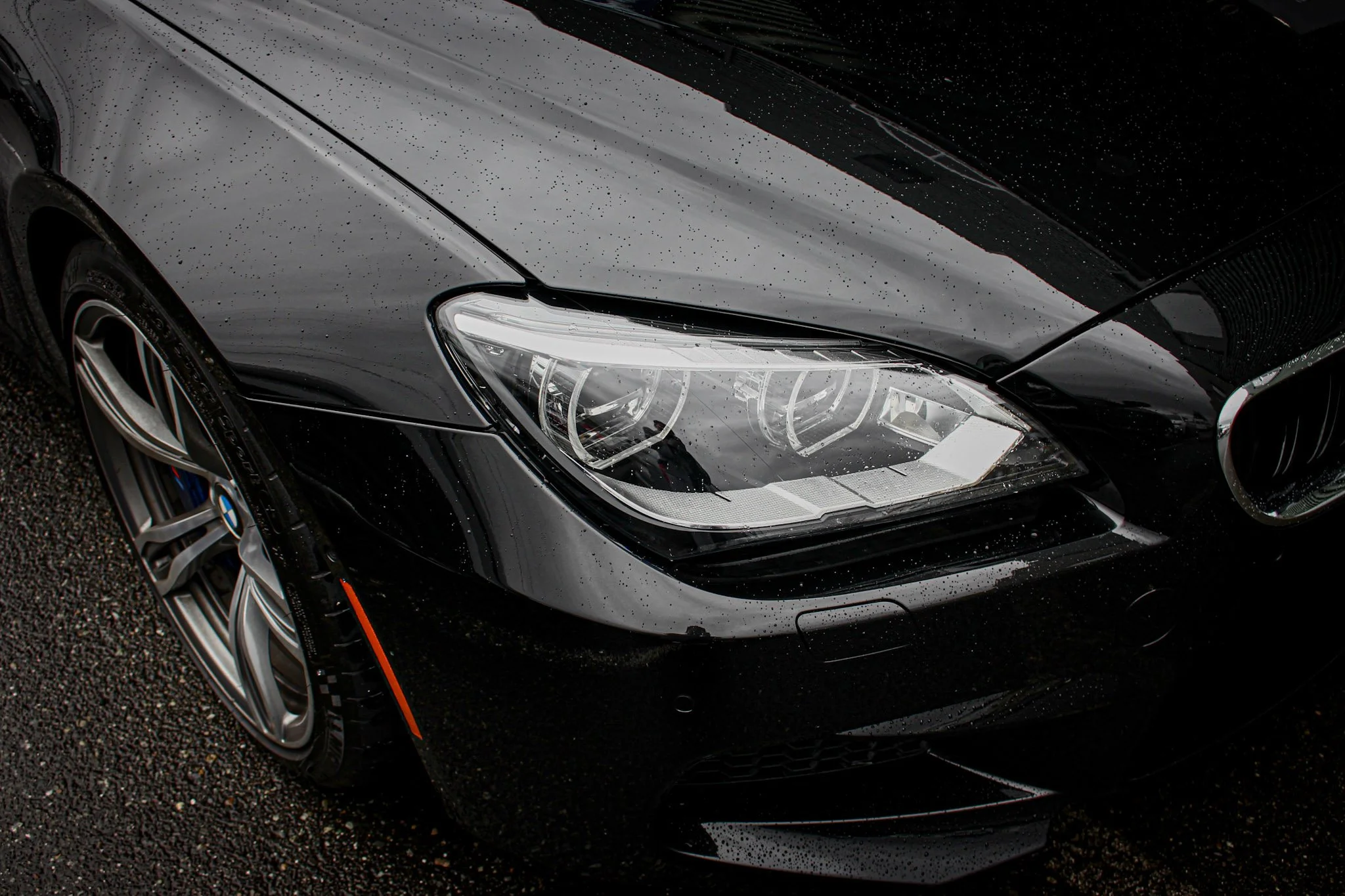 Close-up of a black luxury car with water droplets on the hood and headlight, parked on a wet surface.