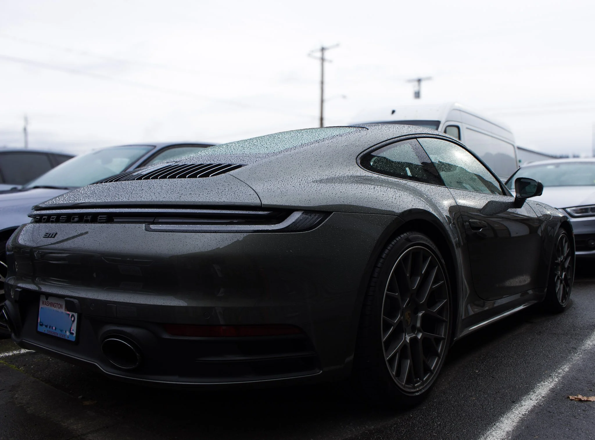 Green Porsche 911 parked in a lot with raindrops on the car and other vehicles in the background under a cloudy sky.