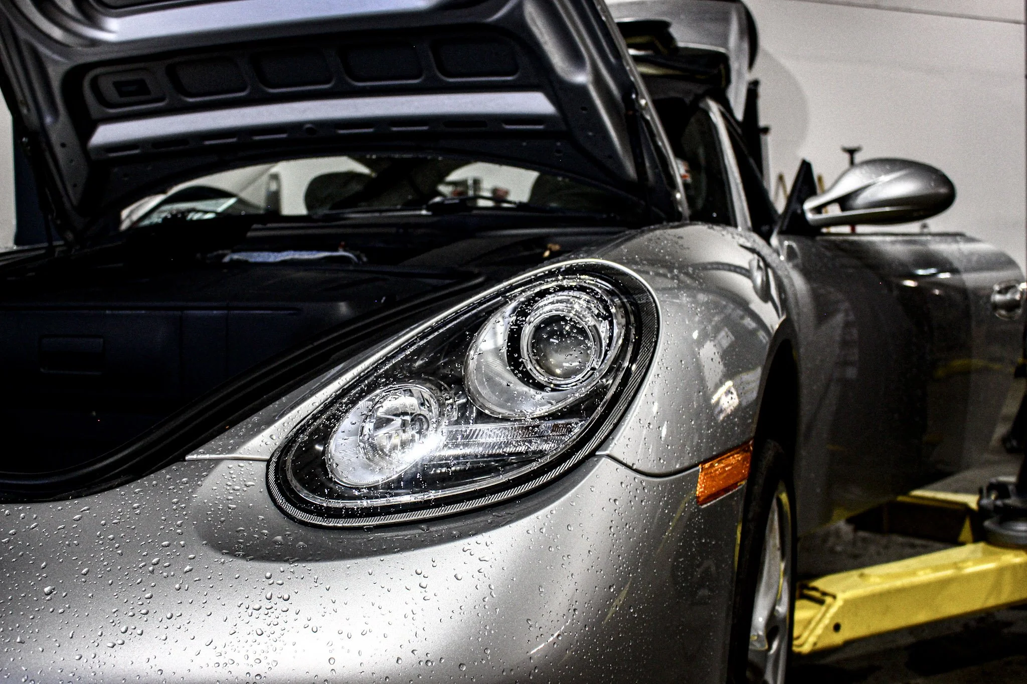 A silver car with water droplets on its surface, parked indoors, with its hood open in a garage or workshop setting.