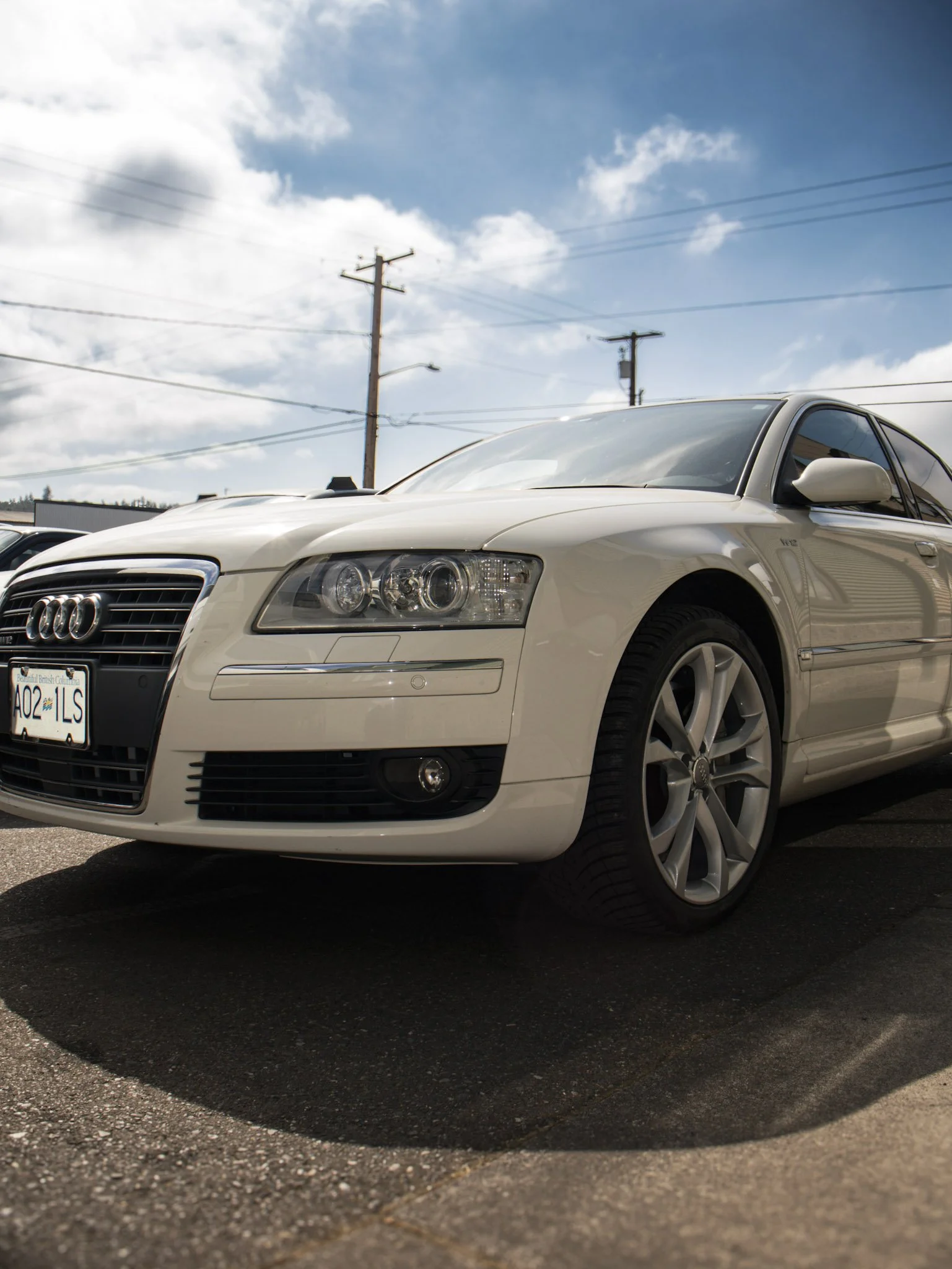 A white Audi sedan parked on a paved surface with power lines and a blue sky with clouds overhead.