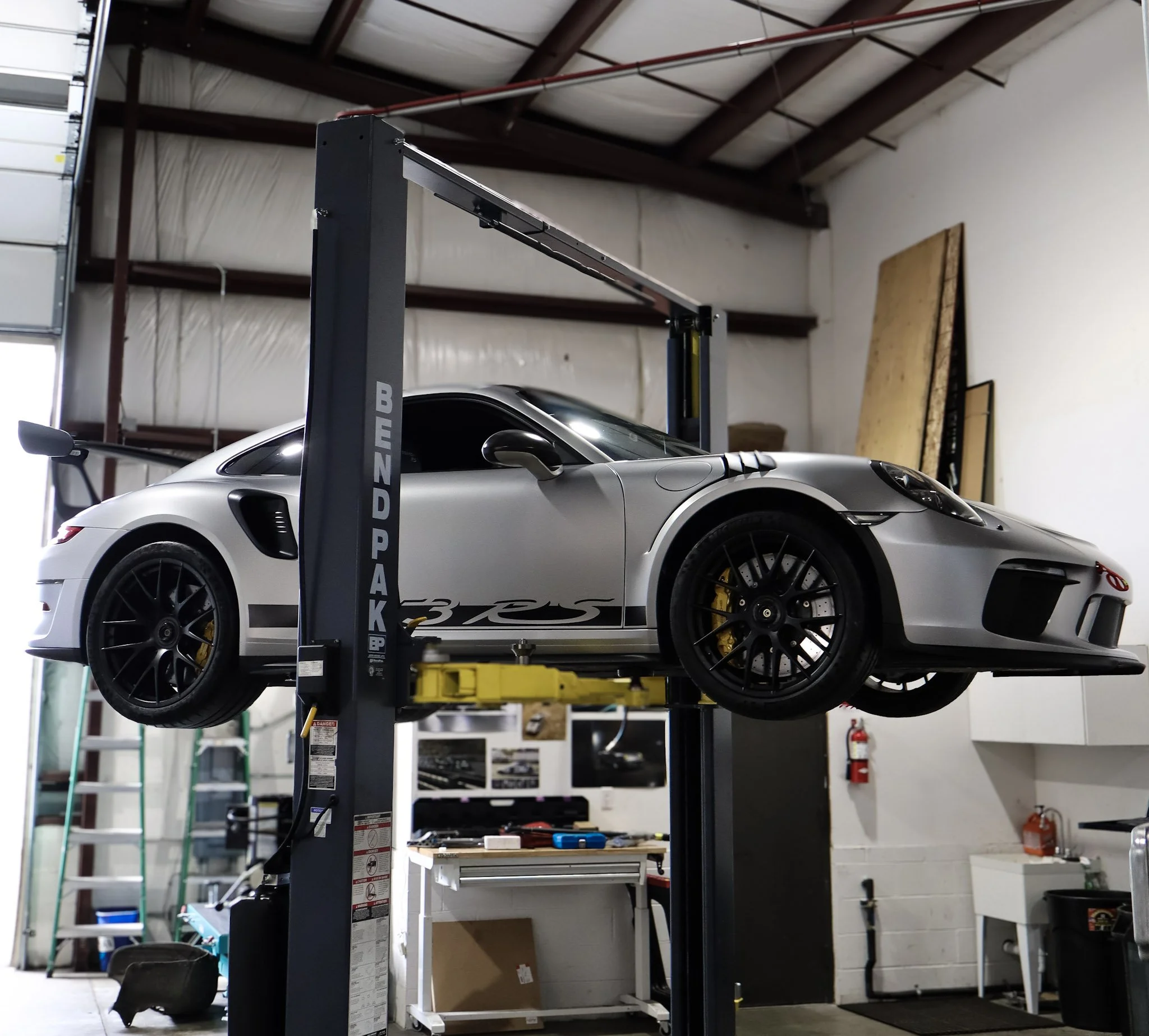 A silver sports car is elevated on a black vehicle lift inside an industrial or warehouse space with high ceilings and exposed beams.