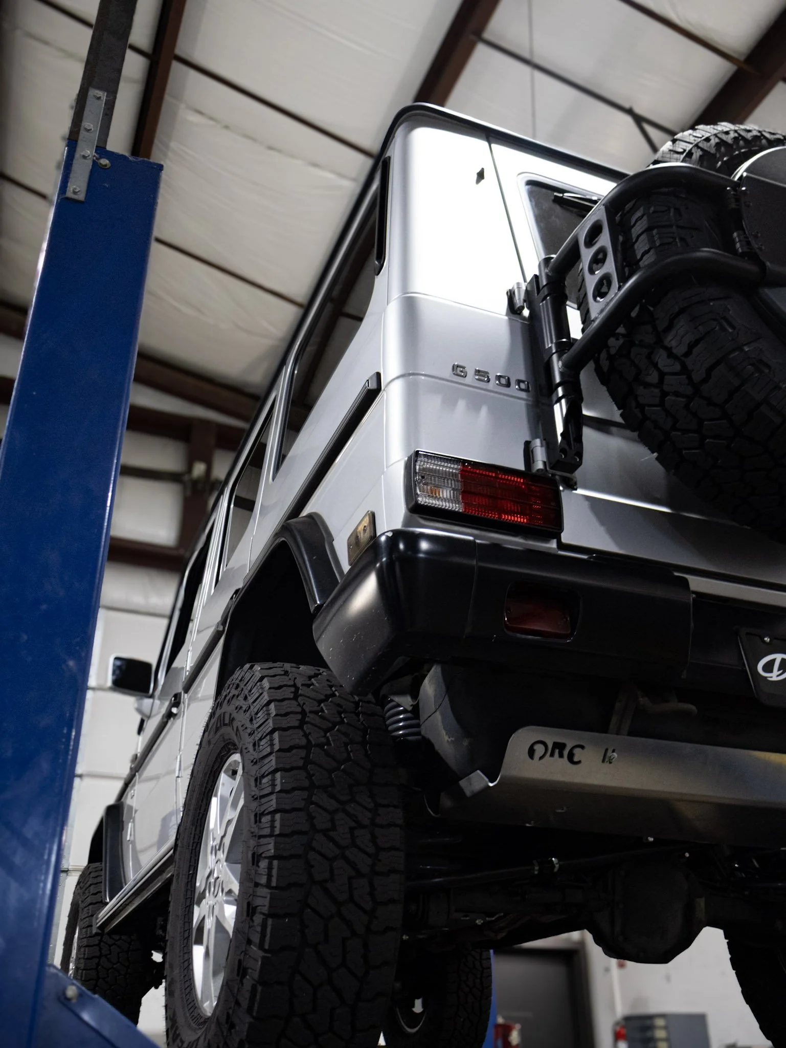 The image shows the back of a white Mercedes-Benz G-Class vehicle with large off-road tires, parked inside a warehouse or garage. The vehicle's rear lights, spare tire, and a portion of the vehicle's side and bumper are visible.