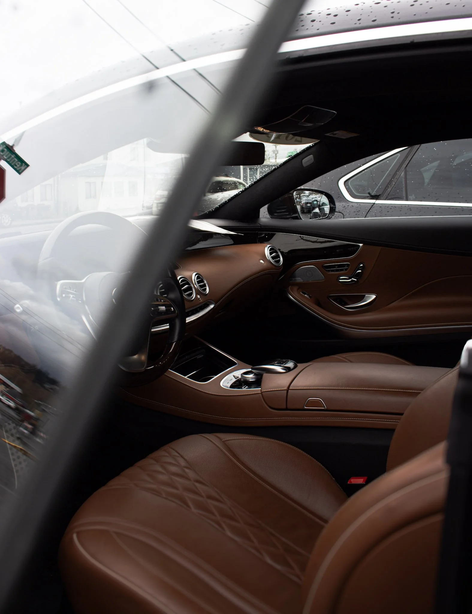 The interior of a luxury car showing tan leather seats, a brown dashboard, and several air vents. Rain droplets are visible on the outside windows.