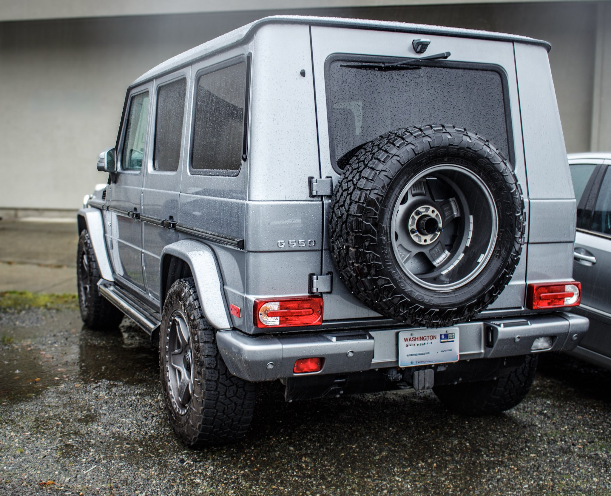 Gray Mercedes-Benz G550 SUV parked on a wet, gravel surface with a spare tire mounted on the rear, wet with rain.