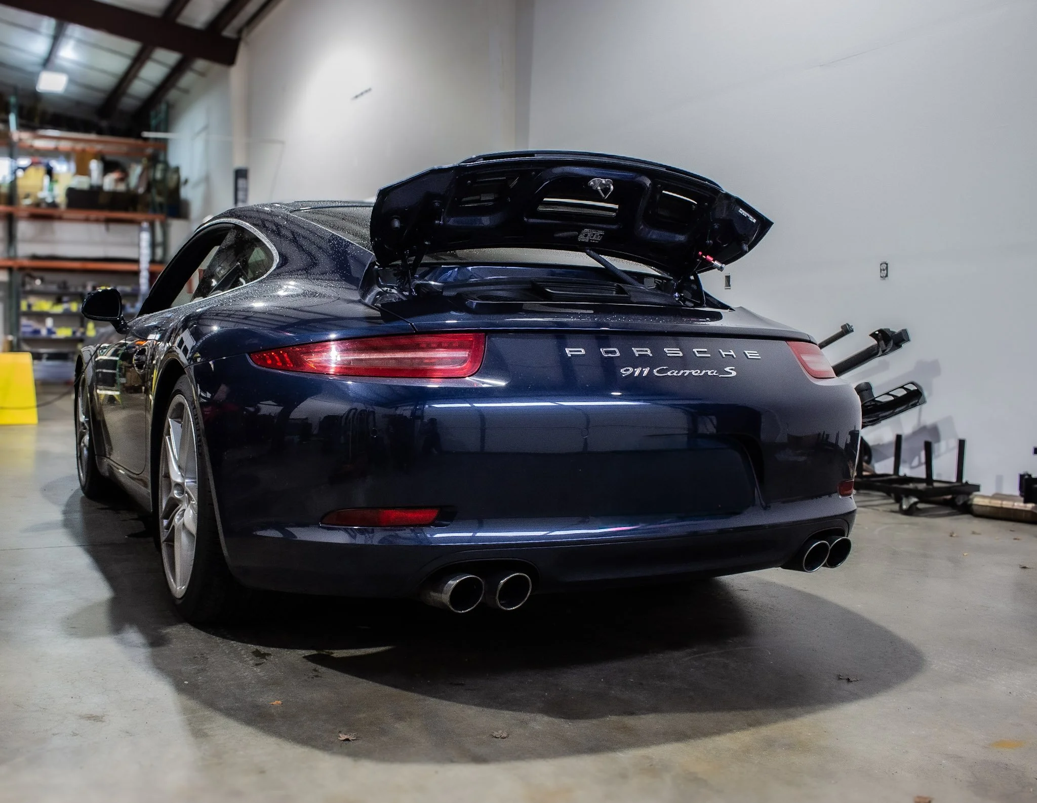 A dark blue Porsche 911 Carrera S in a garage with the rear hatch open, showing the dual exhaust pipes and the garage interior with shelves and equipment.