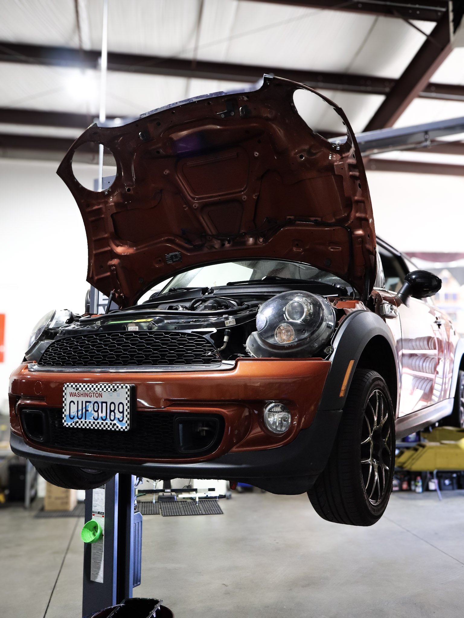 A red Mini Cooper car is elevated on a hydraulic lift in a garage, with its hood open, exposing the engine compartment. The garage has a concrete floor and various tools and equipment in the background.