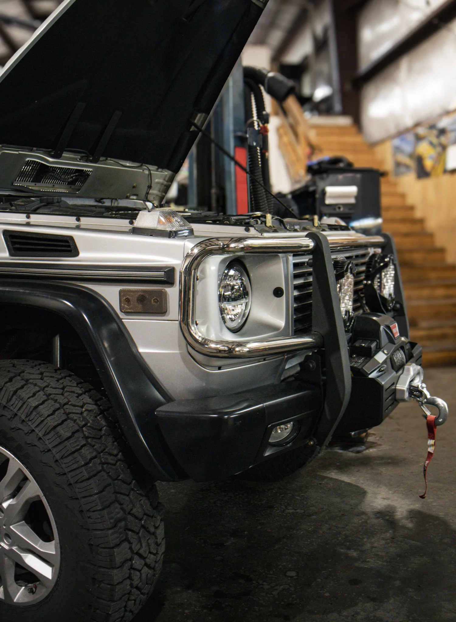 Close-up of the front of a silver off-road vehicle with large tires, a bumper guard, and an open hood, parked on a dark pavement in a garage with wooden walls.
