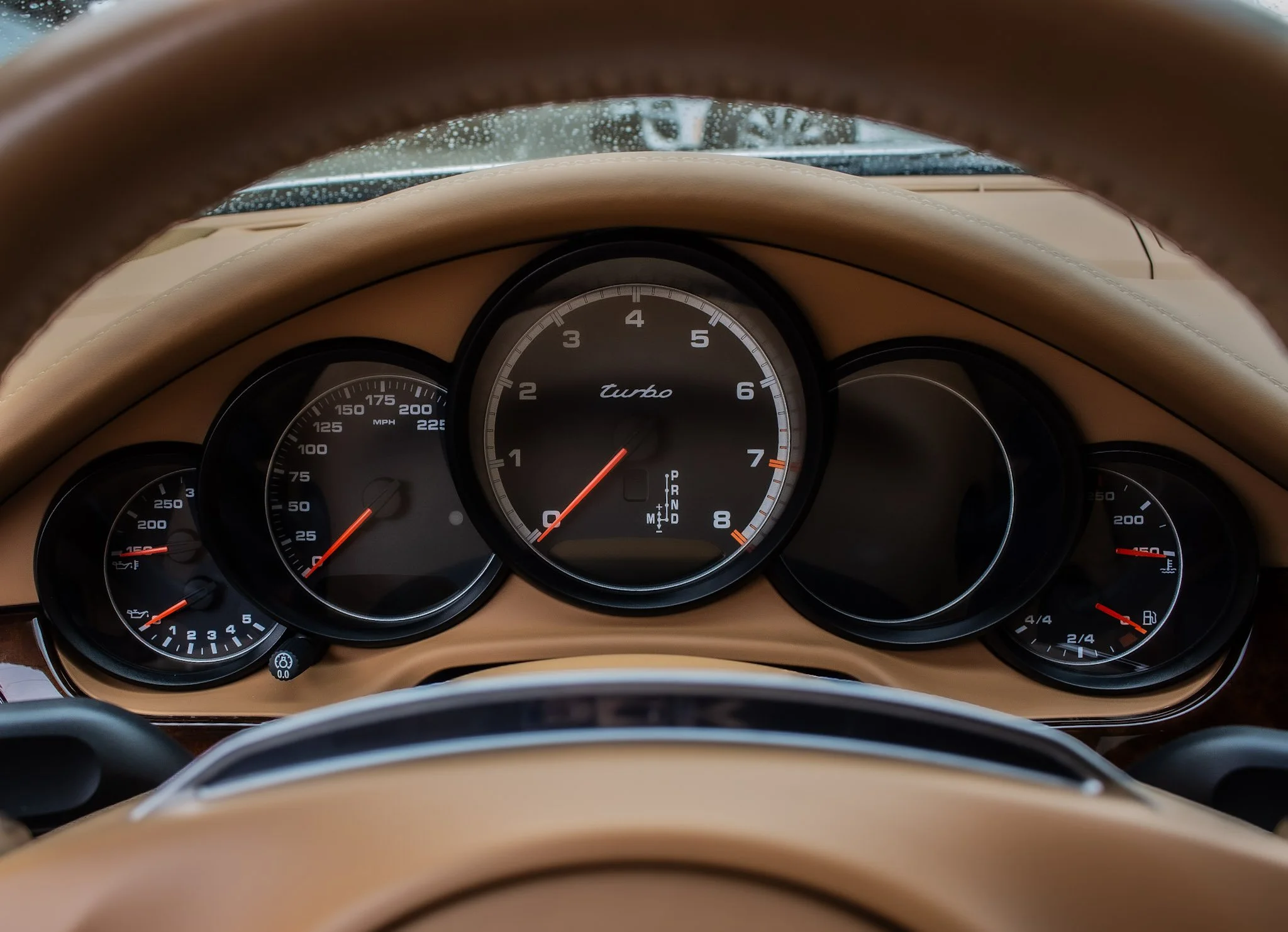View from inside a car showing instrument cluster with speedometer, tachometer, fuel gauge, and temperature gauge, with steering wheel and dashboard in beige color.
