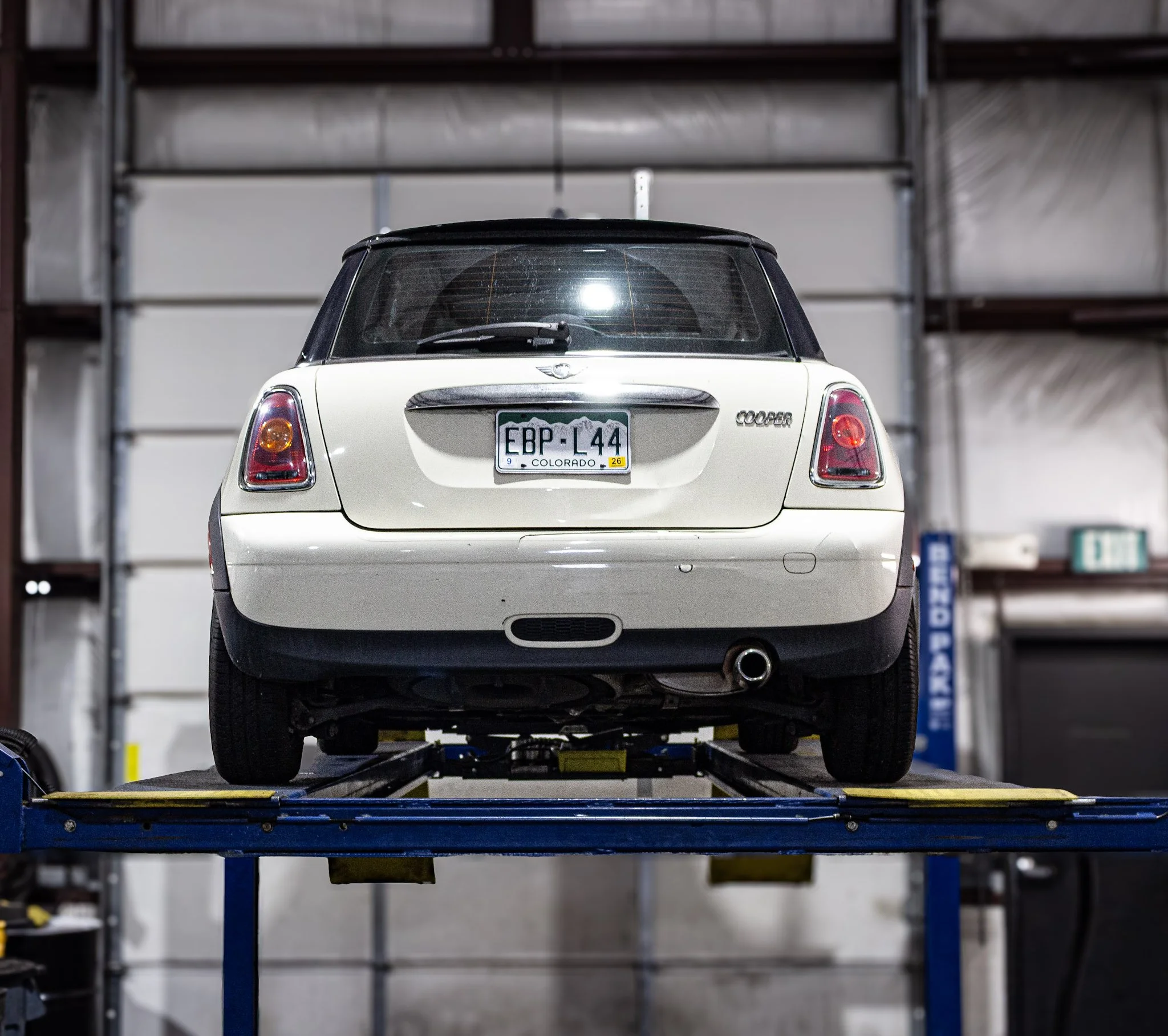 A white Mini Cooper car on a hydraulic lift inside a garage or workshop. The garage has a metal shelving unit in the background. The license plate reads 'EBP-L44' from Colorado.
