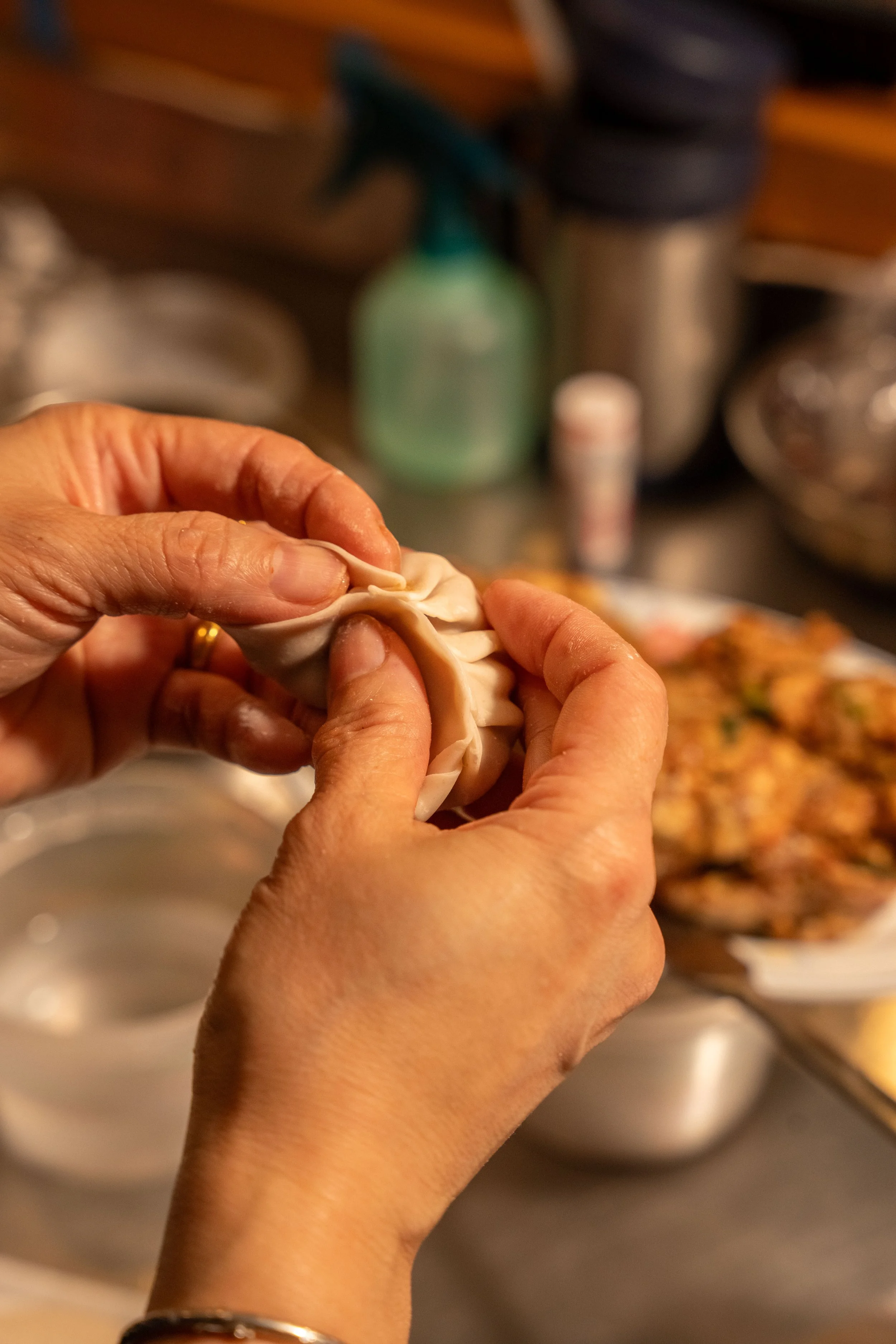 Hands folding a dumpling in a kitchen with various containers and food in the background.