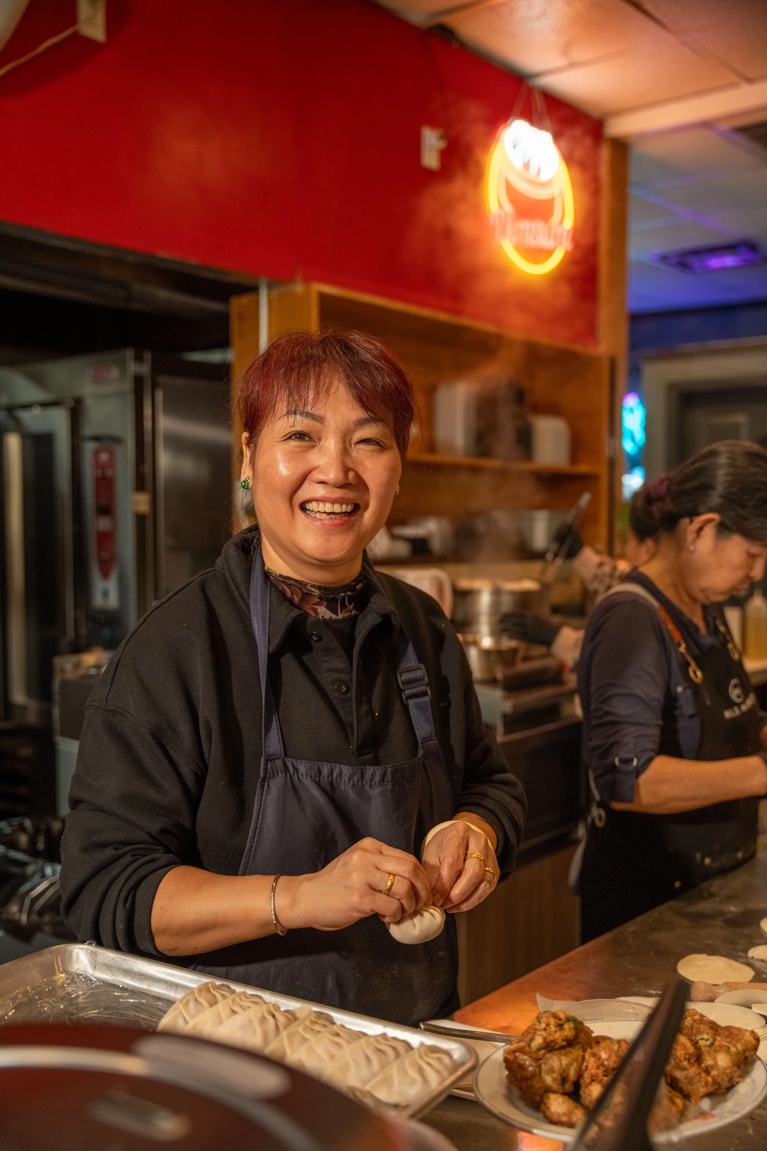 A woman in a black apron is smiling while preparing dumplings in a restaurant kitchen, with another person working behind her and a neon sign on the wall.