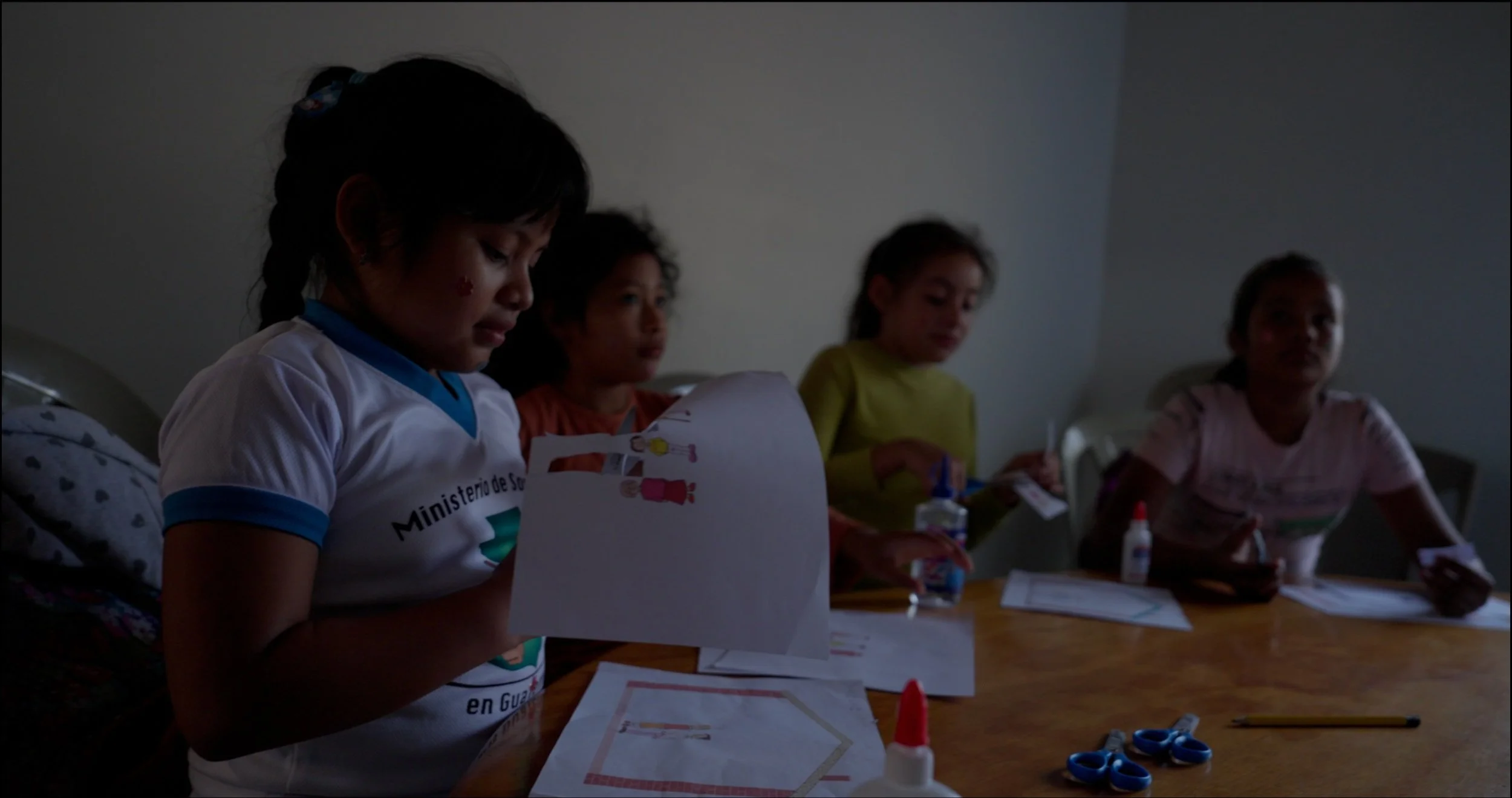 Four young girls seated at a table engaged in arts and crafts activities, with craft supplies like scissors, glue, and paper in front of them, in a dimly lit room.