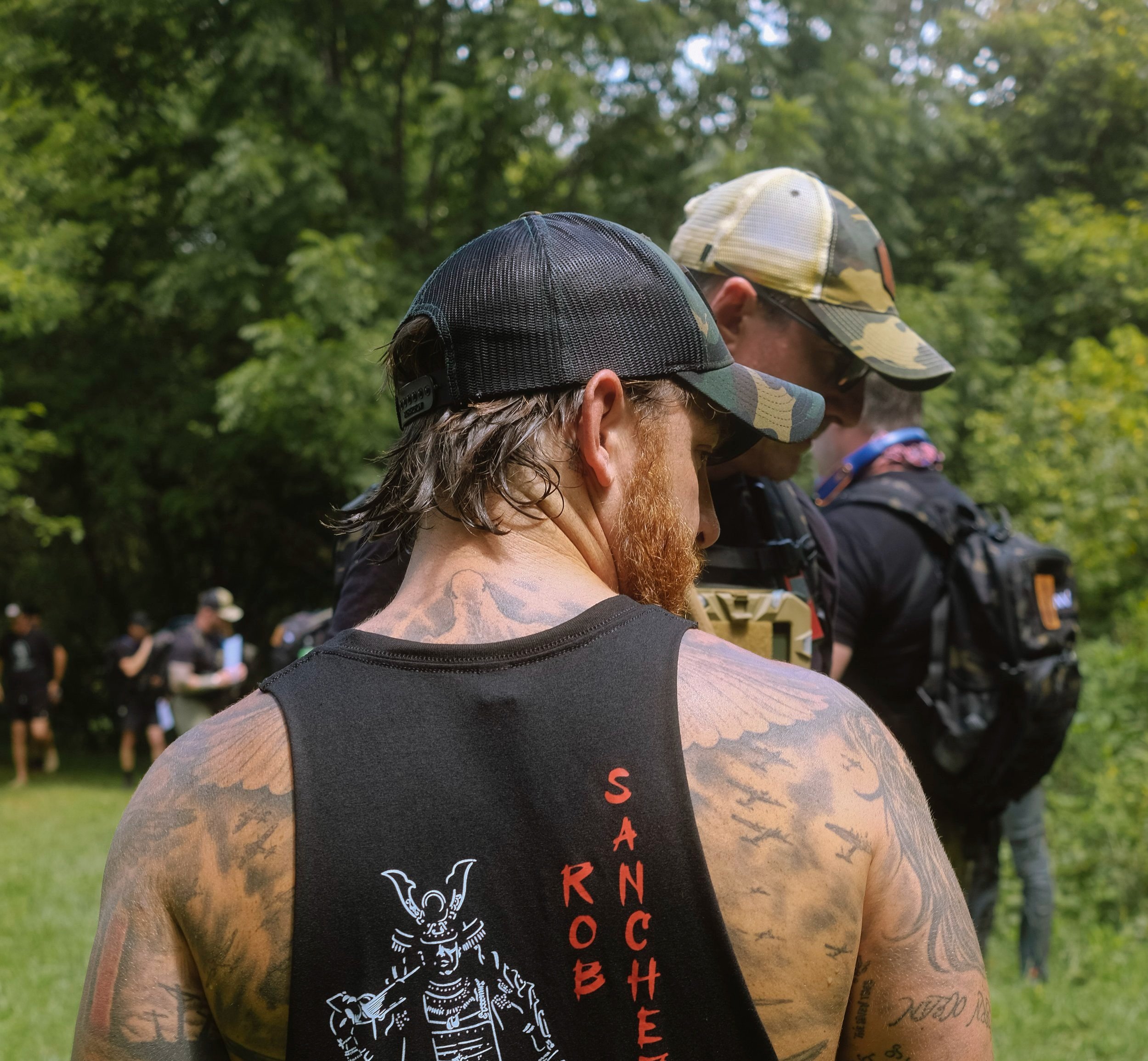 Two men in outdoor setting, wearing caps and backpacks, one with tattoos and a graphic tank top.