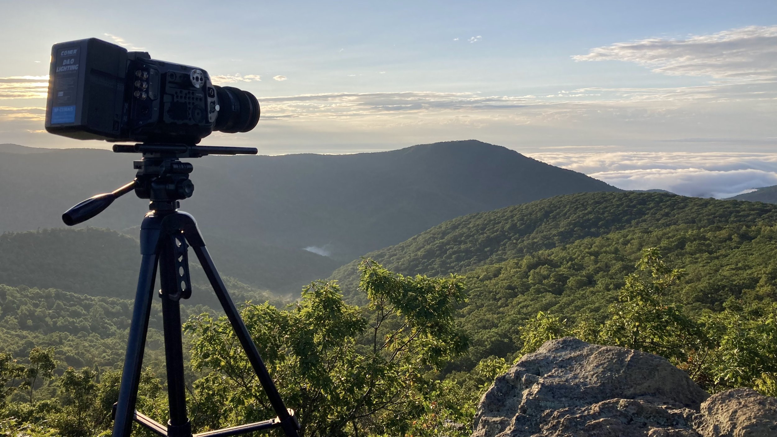 Camera on a tripod facing a scenic mountain landscape with cloudy sky at sunrise.