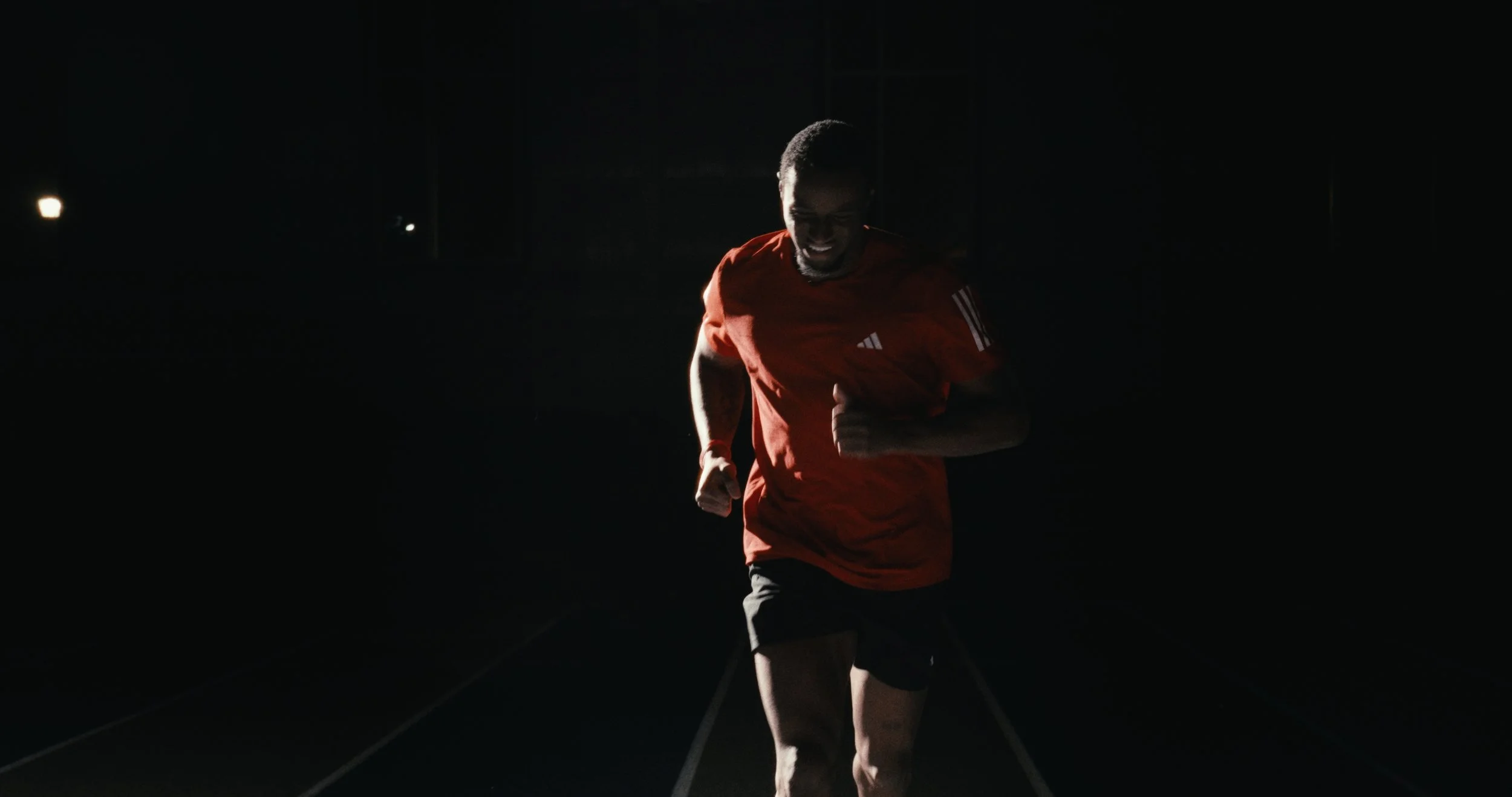 A man running indoors in a dark room, illuminated by a light source behind him, wearing a red sports shirt and black shorts.