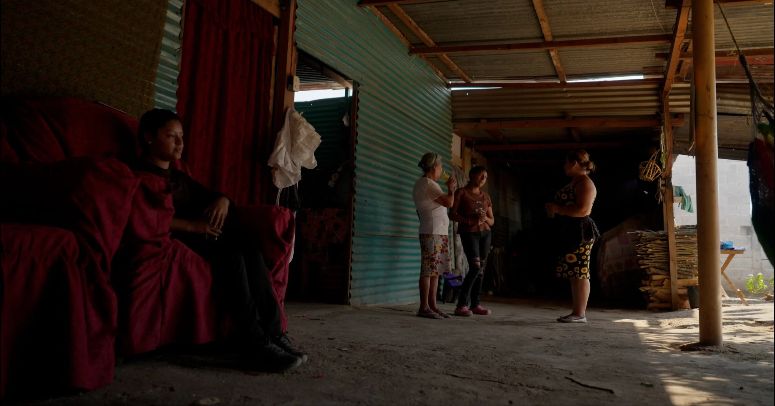 Four women are standing and talking inside a rustic, open-sided structure with a dirt floor. One woman is sitting on a red couch, and soft sunlight is coming in from outside.