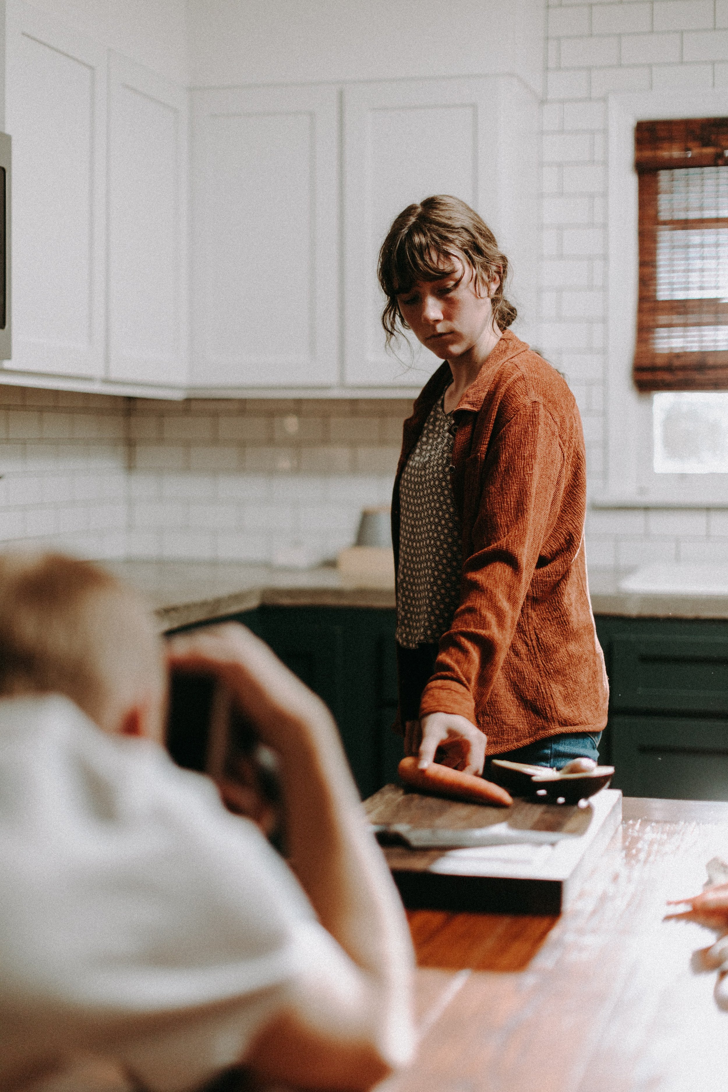 Woman in a kitchen holding a frying pan, gazing intently, with another person seated in foreground.