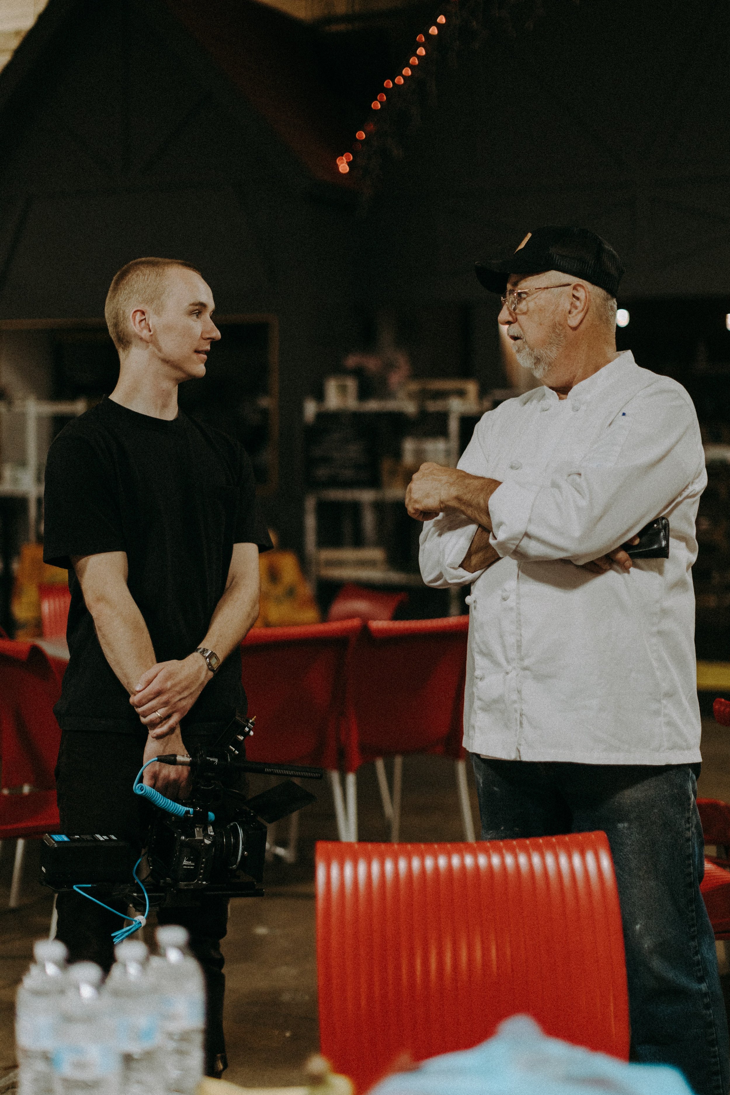 Two men having a conversation in an indoor setting, one holding a camera, the other wearing a chef's coat and hat.