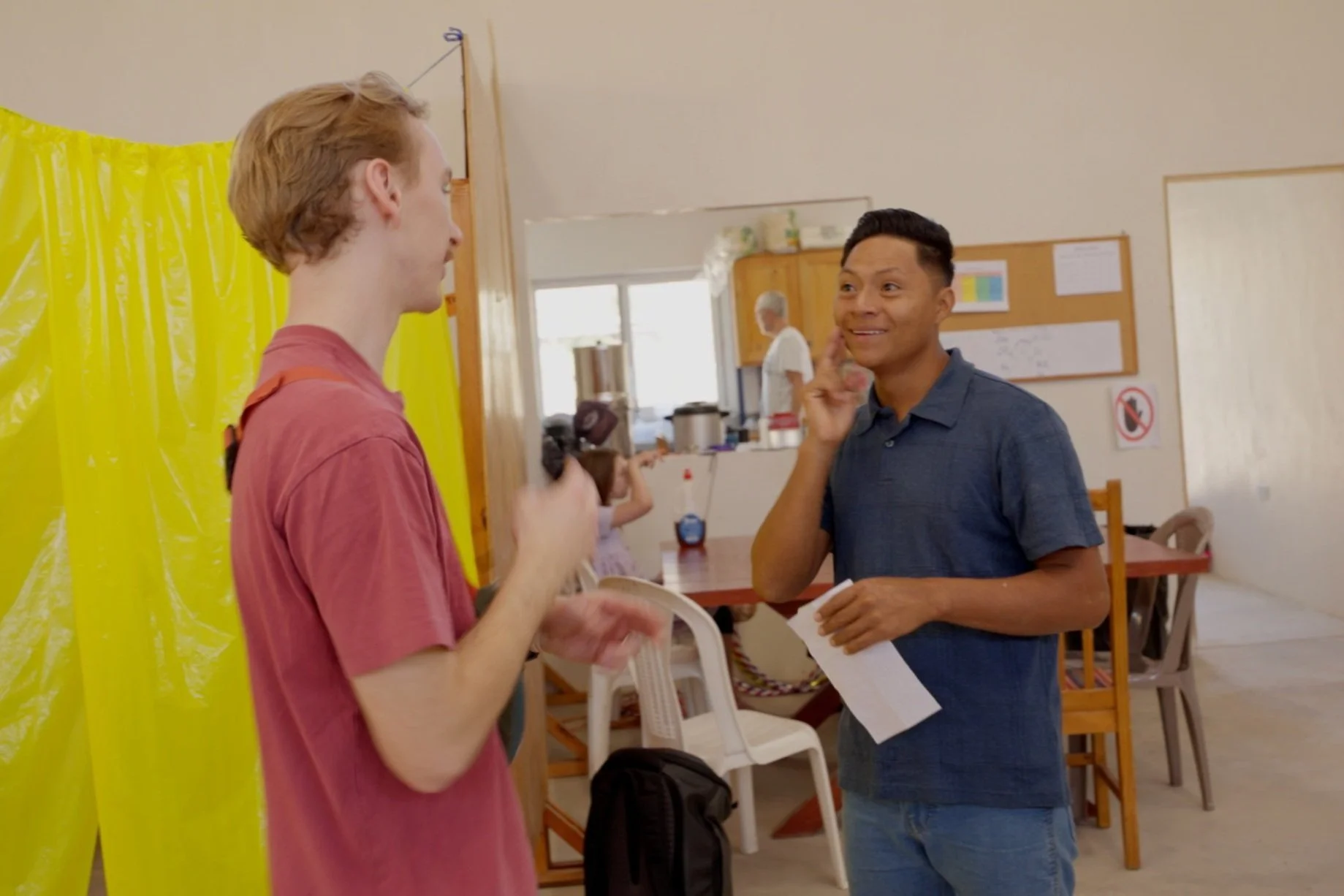 Two young men having a conversation in sign language indoors near a yellow plastic curtain, with a woman in white in the background, a table, chairs, and a bulletin board on the wall.