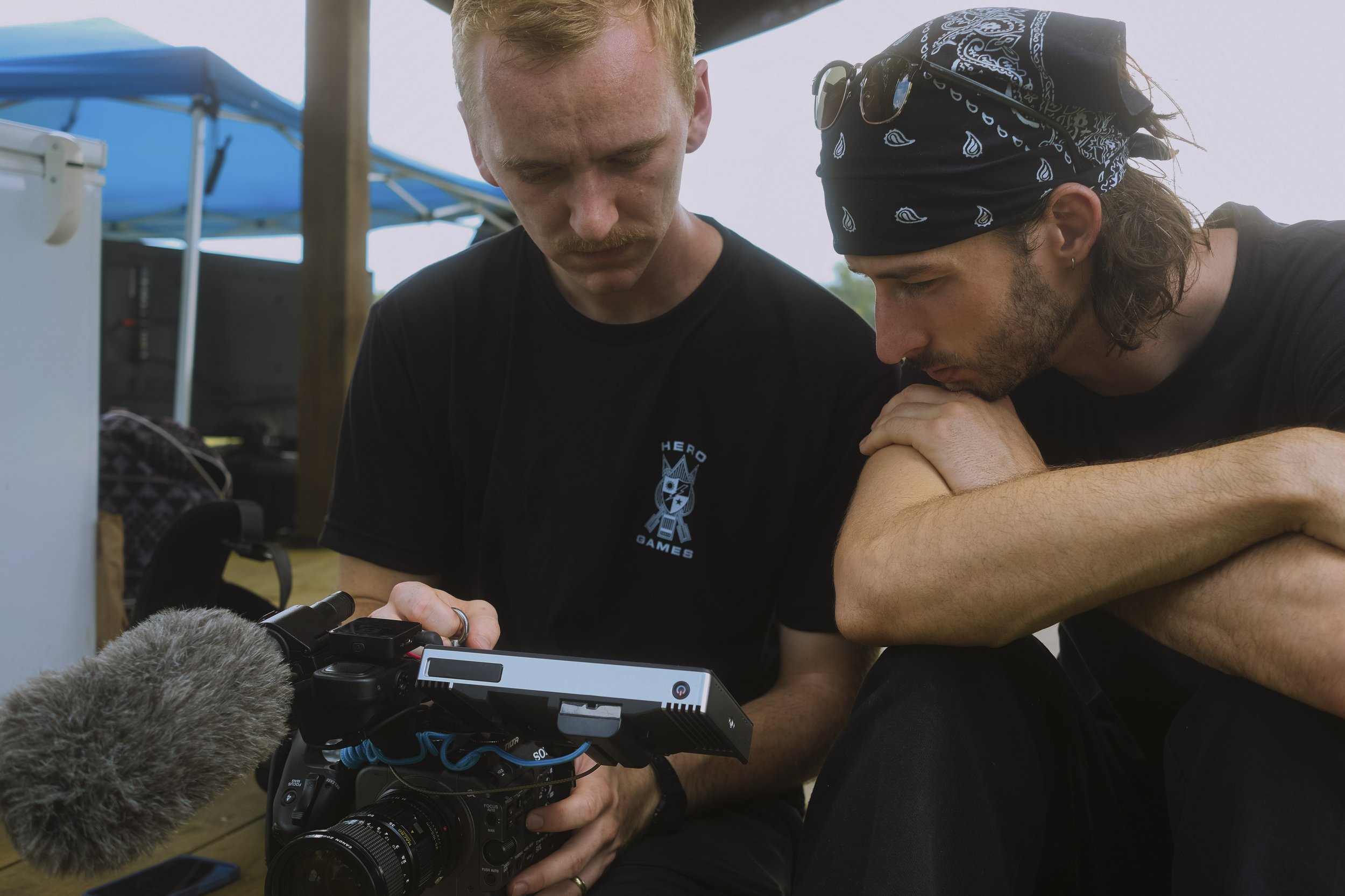 Two men looking at a camera with a microphone outdoors under a canopy.